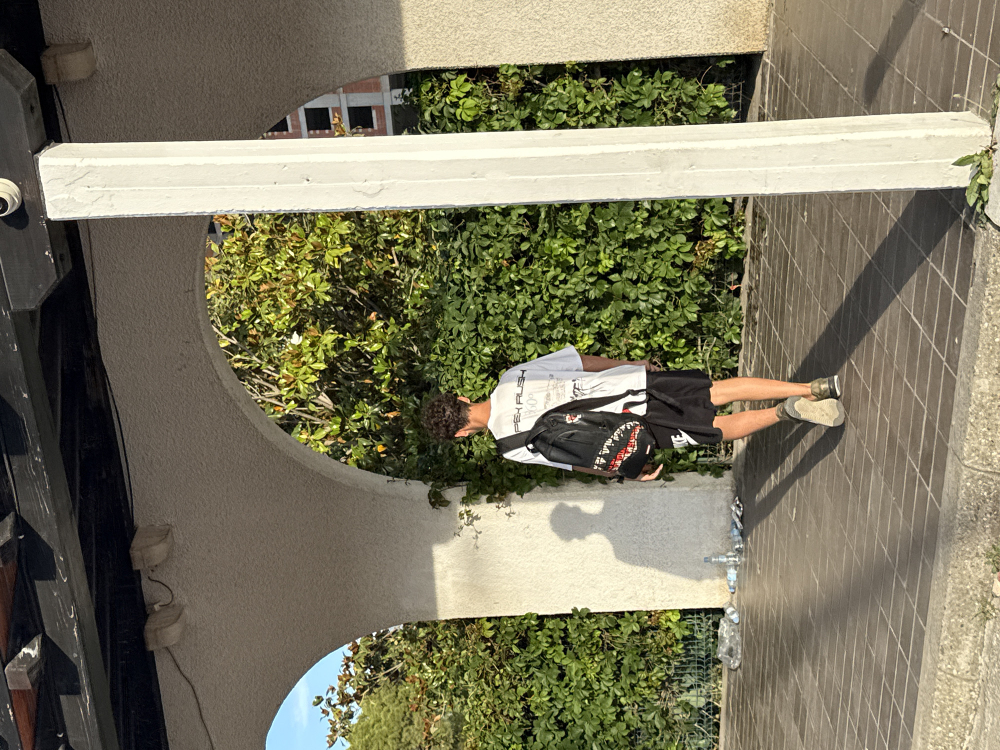 The image captures a full-length, upright view of a young male standing in an arched outdoor walkway, likely in Budva, Montenegro. The person is seen from behind, looking away from the camera towards a lush green area. He has dark, curly hair and is wearing a white t-shirt, black shorts, and khaki-colored sneakers. A black backpack with a prominent shark teeth design is slung over his shoulders. Visible text on his t-shirt includes "PEK PUSH" and "1980".
The setting appears to be a sunny day, indicated by the bright lighting and long shadows cast on the tiled floor, suggesting either morning or late afternoon. The walkway features light-colored, textured archways and columns, contrasting with a dark, tiled wall/floor on the right side of the frame. Abundant green foliage, possibly vines or bushes, is visible through the archways and around the structures, with a glimpse of a distant building through the leaves.
Several discarded plastic bottles are scattered on the tiled ground near the person's feet. A small security camera is mounted on a dark pillar to the left, near the start of the archway. The overall scene conveys a casual, outdoor moment within an architectural space surrounded by nature.