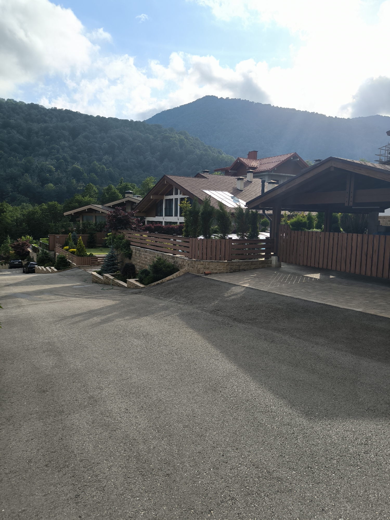 The image shows a view of several modern houses with wooden accents, featuring sloped, dark-tiled roofs and large windows. The houses are situated on a hillside surrounded by lush green trees and mountains in the background, under a partly cloudy sky with sunlight filtering through. A paved road with a shadow of trees crosses the foreground, leading up to the properties. The properties are enclosed by wooden fences and stone retaining walls. Two black cars are parked on the side of the road. The scene appears to be in a residential area in Krasnaya Polyana, Russia, known for its mountainous landscape and resorts.