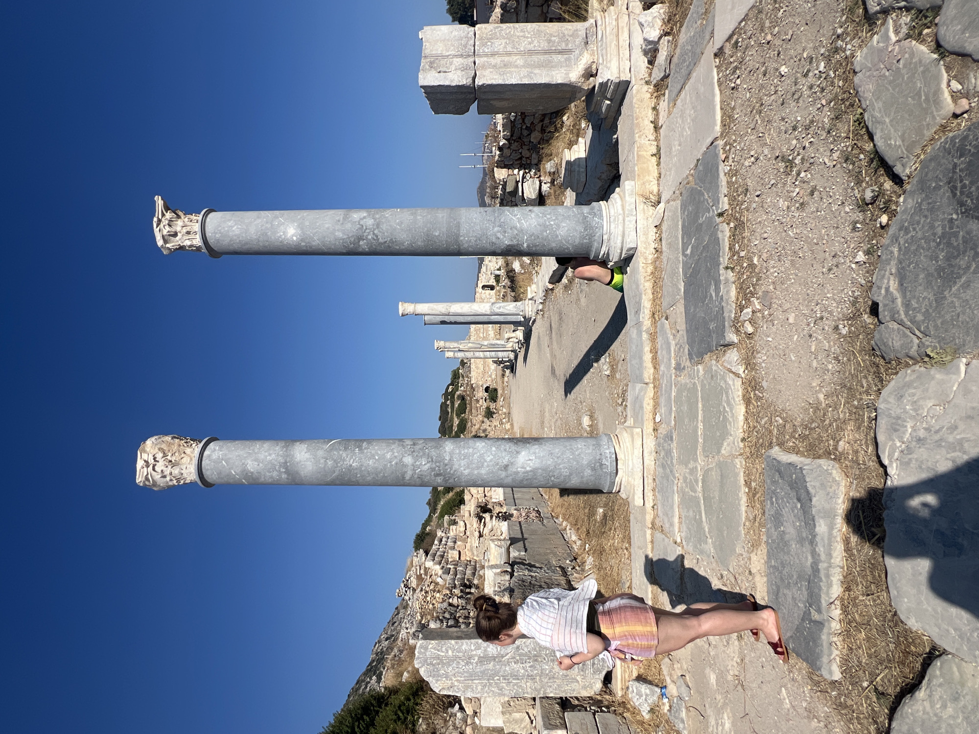 The image shows an outdoor scene at an archaeological site with ancient ruins under a clear blue sky. In the foreground, a woman stands to the left, facing away from the camera and looking towards the ruins. She is wearing a white and striped top and a colorful striped skirt. Behind her, to the right, a second person is partially visible, crouching behind a large stone column. This person is wearing bright green shorts.
The ruins consist of stone columns, some intact and some broken, and stone blocks scattered on the ground. A paved pathway made of large stone slabs runs through the site. In the background, more ruins are visible, along with a hillside with sparse vegetation. The overall environment suggests a hot, sunny day in a historical location.
