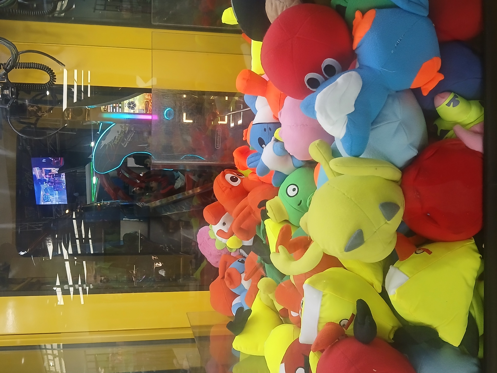 This vertical shot captures the interior of a claw machine filled with a colorful assortment of plush toys. The subjects are numerous stuffed animals, predominantly in shades of red, blue, green, and yellow. Notable among them are a large red octopus-like plush with prominent eyes, several blue duck-like or bird-like plushies with orange features, and multiple yellow and red plush figures resembling characters from the "Angry Birds" franchise, including the yellow 'Chuck' bird and red 'Red' bird, as well as green pig characters.
The claw machine's yellow casing is visible at the top. The glass front creates significant reflections, revealing elements of the surrounding arcade. Reflected details include a monitor displaying gameplay, vibrant neon lighting in pink, blue, and green from other arcade games, and partial text. The word "Nintendo" is clearly visible in one reflection, likely from an adjacent machine. Another reflection shows the partial text "Kart Fighter Blast," suggesting a racing game. The coiled wire and parts of the claw mechanism itself are visible in the upper left.
The scene is set indoors, within what appears to be an arcade or entertainment center in Guayana City, Venezuela, characterized by the type of game and the reflections of other electronic amusements. No people are directly visible, but the setup implies an active entertainment environment. The bright colors of the toys and the reflected arcade lights suggest a lively atmosphere.