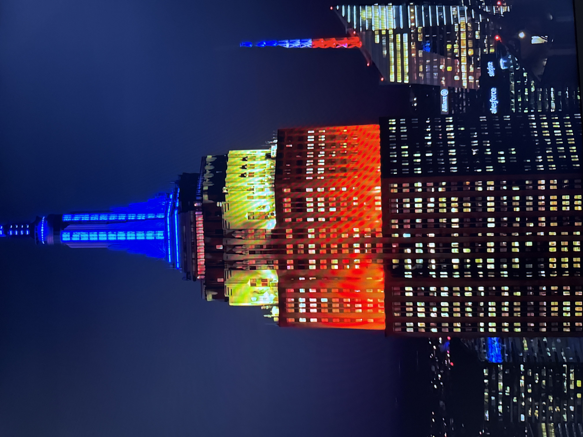 The image shows the Empire State Building in New York City illuminated at night with blue and yellow lights on its spire and upper floors, and red lights on its lower illuminated floors. This illumination is reminiscent of the Ukrainian flag. To the right of the Empire State Building, another skyscraper is visible, also lit with blue and red lights. The foreground and background are filled with the dark night sky and the distant city lights, indicating an urban environment. The image does not depict Burgas, Bulgaria.