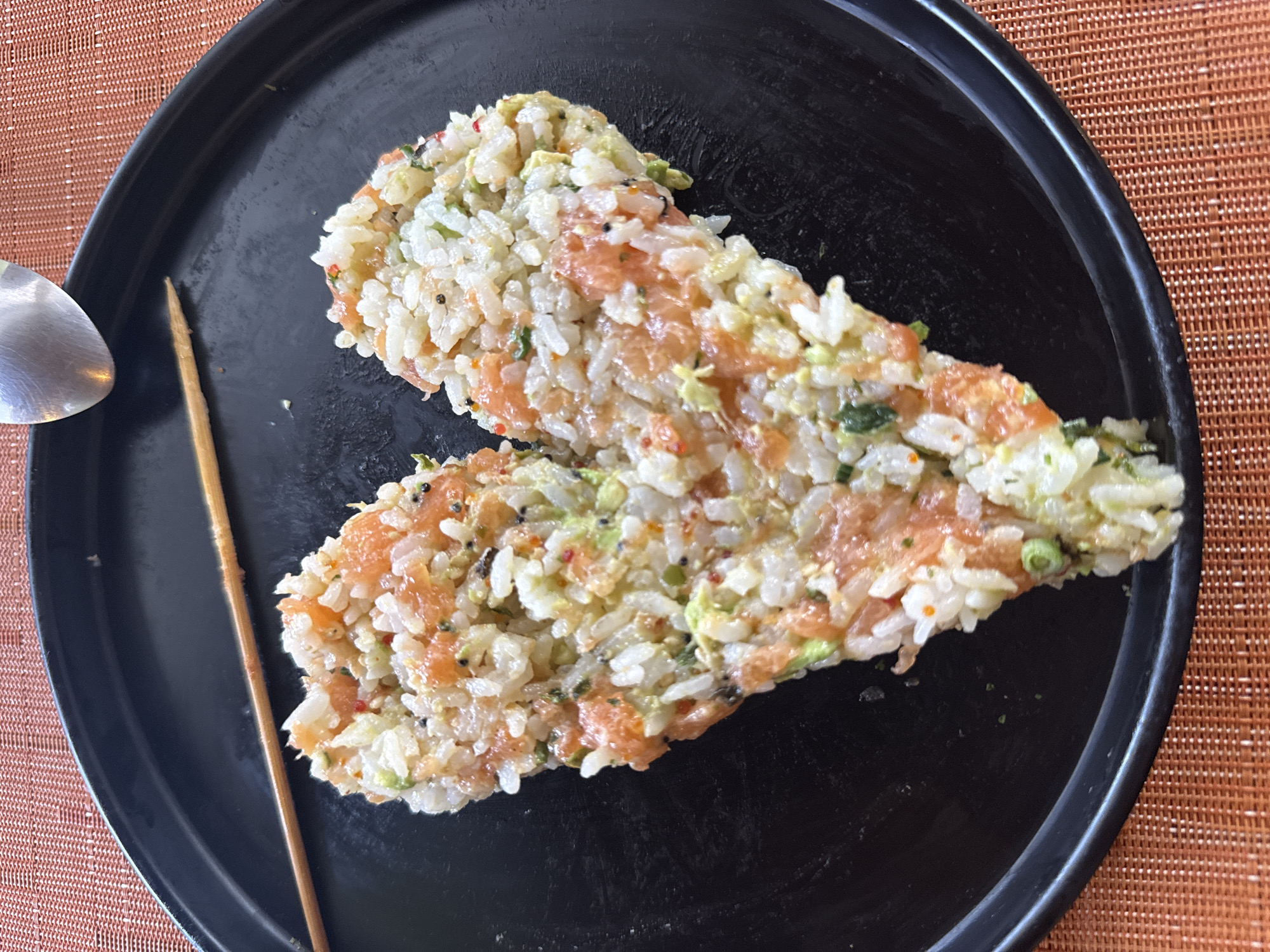 A black plate holds two irregularly shaped portions of food. The food appears to be a mixture of rice and salmon, with specks of green and black visible throughout. A metal spoon is visible on the left side of the plate, and a thin, brown chopstick rests beside the food. The plate sits on a surface with a woven texture in shades of brown and orange. The photo was taken in San Bernardino Tlaxcalancingo, Mexico.