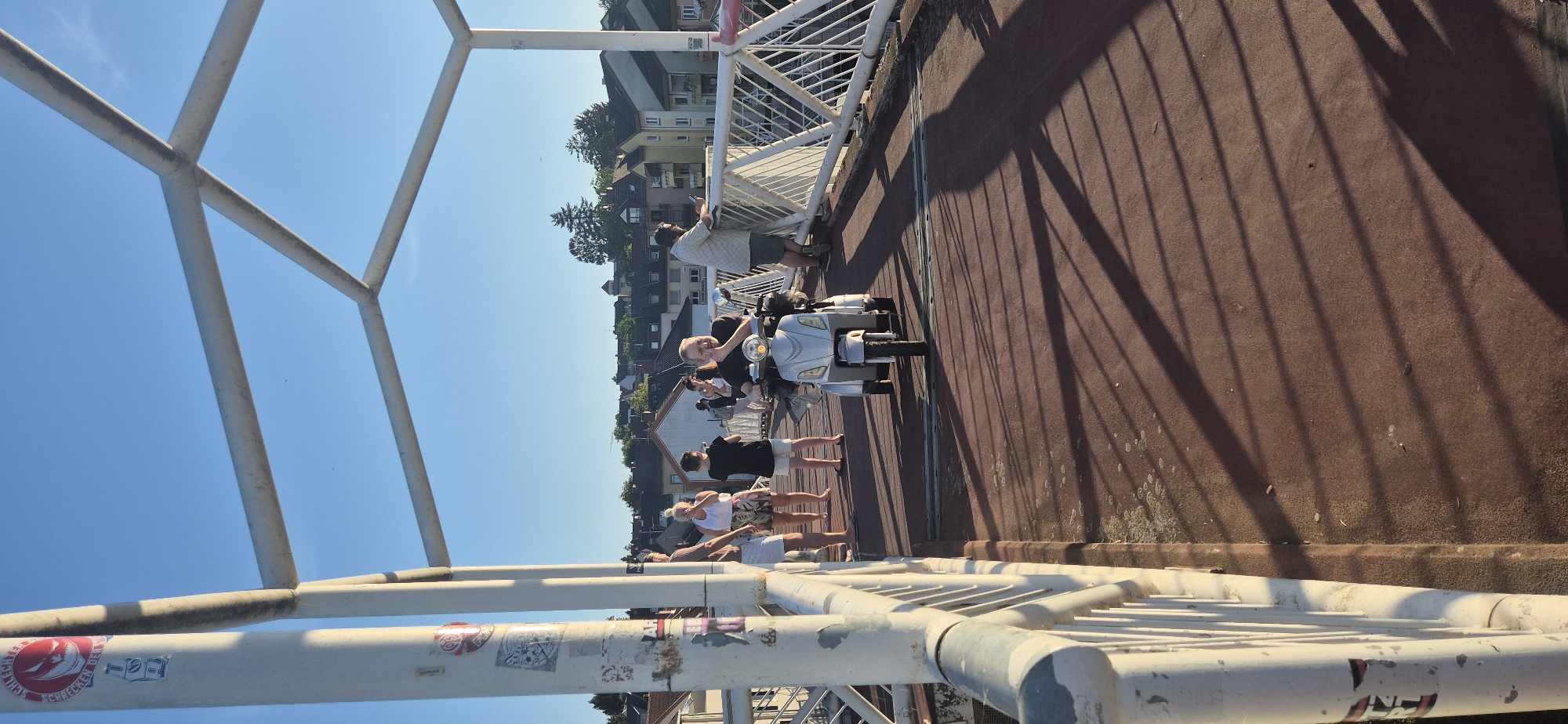 The image depicts a sunny day scene on a pedestrian bridge in Mainz, Germany. The bridge has a red-brown surface and a white metal railing. Several people are present on the bridge. In the foreground, a man is riding a silver motorized scooter. Further back, a group of people are standing, some conversing, while others are looking around. The background shows a cityscape with buildings and trees under a clear blue sky. The lighting suggests it is late afternoon or early evening, with long shadows cast by the bridge's structure and the people. The bridge appears to be a modern construction with metal supports and railings.