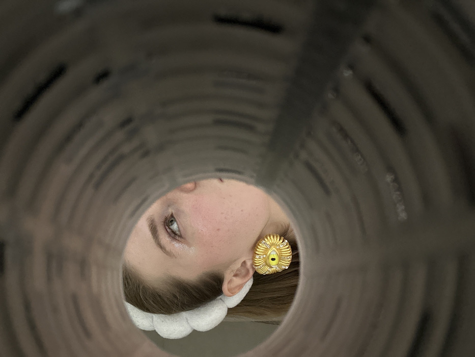 A face peers through the circular opening of a gray tube. The visible eye is light with sparse lashes. A golden, textured earring adorned with small diamonds is on the visible ear. A white, fluffy headband sits on the hair. The tube walls are textured, appearing to be a tunnel of printed material. The lighting is soft, casting shadows and highlighting the textures.