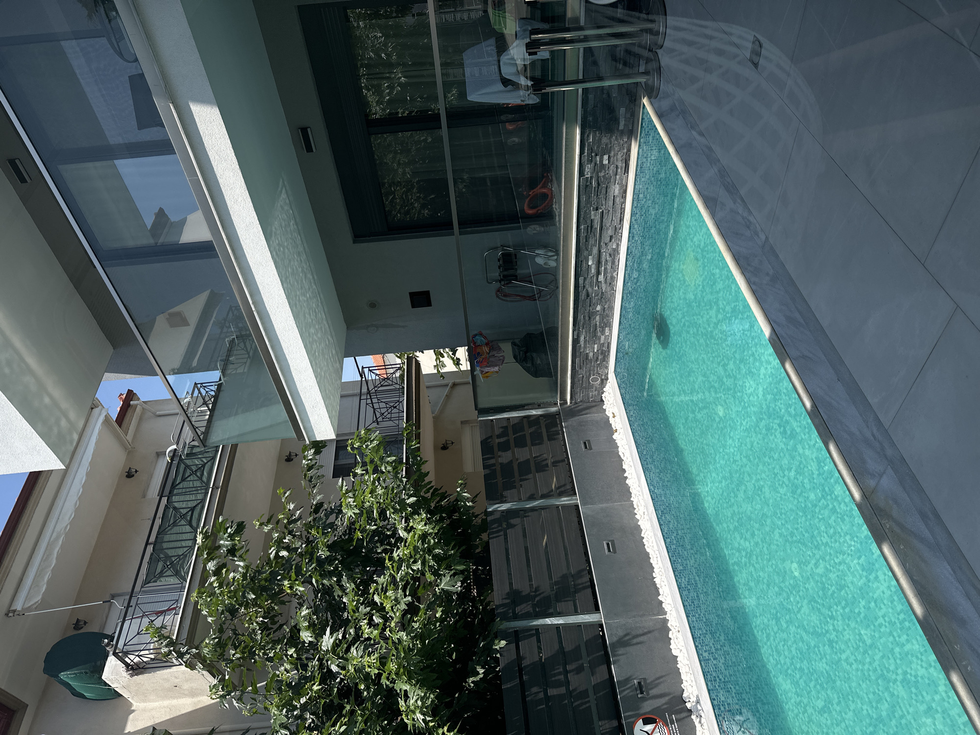 A swimming pool is shown in the foreground, filled with turquoise water and lined with white pebbles. Behind the pool is a modern building with glass railings and balconies. To the left of the pool, a dark wooden fence separates the pool area from a lush green tree and the facade of an older building. The image appears to be taken on a sunny day. The location is Ofrynio Beach, Greece.