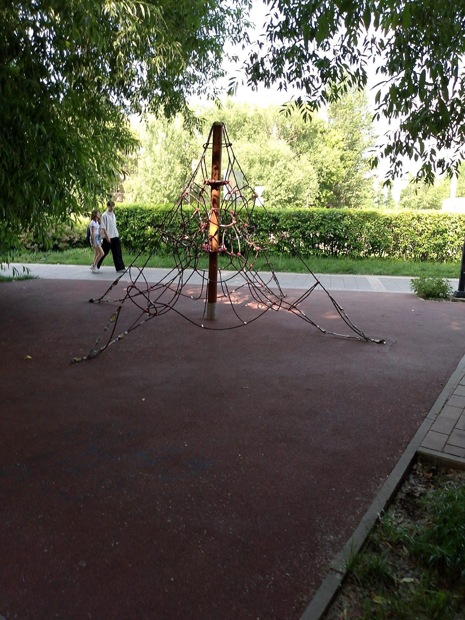 In an outdoor urban park setting, likely in Astana, Kazakhstan, a children's climbing net structure made of ropes and a central wooden pole stands on a dark red rubberized surface. The structure is anchored to the ground with metal fasteners. Two individuals, a young girl and a boy, walk on a paved path adjacent to the climbing net. The girl is wearing a white t-shirt and denim shorts, carrying a backpack. The boy is dressed in a light-colored button-down shirt and dark trousers. Behind the path, a dense green hedge forms a boundary, with trees and foliage visible in the background, suggesting a sunny day. The scene captures a moment of pedestrian activity in a recreational area.