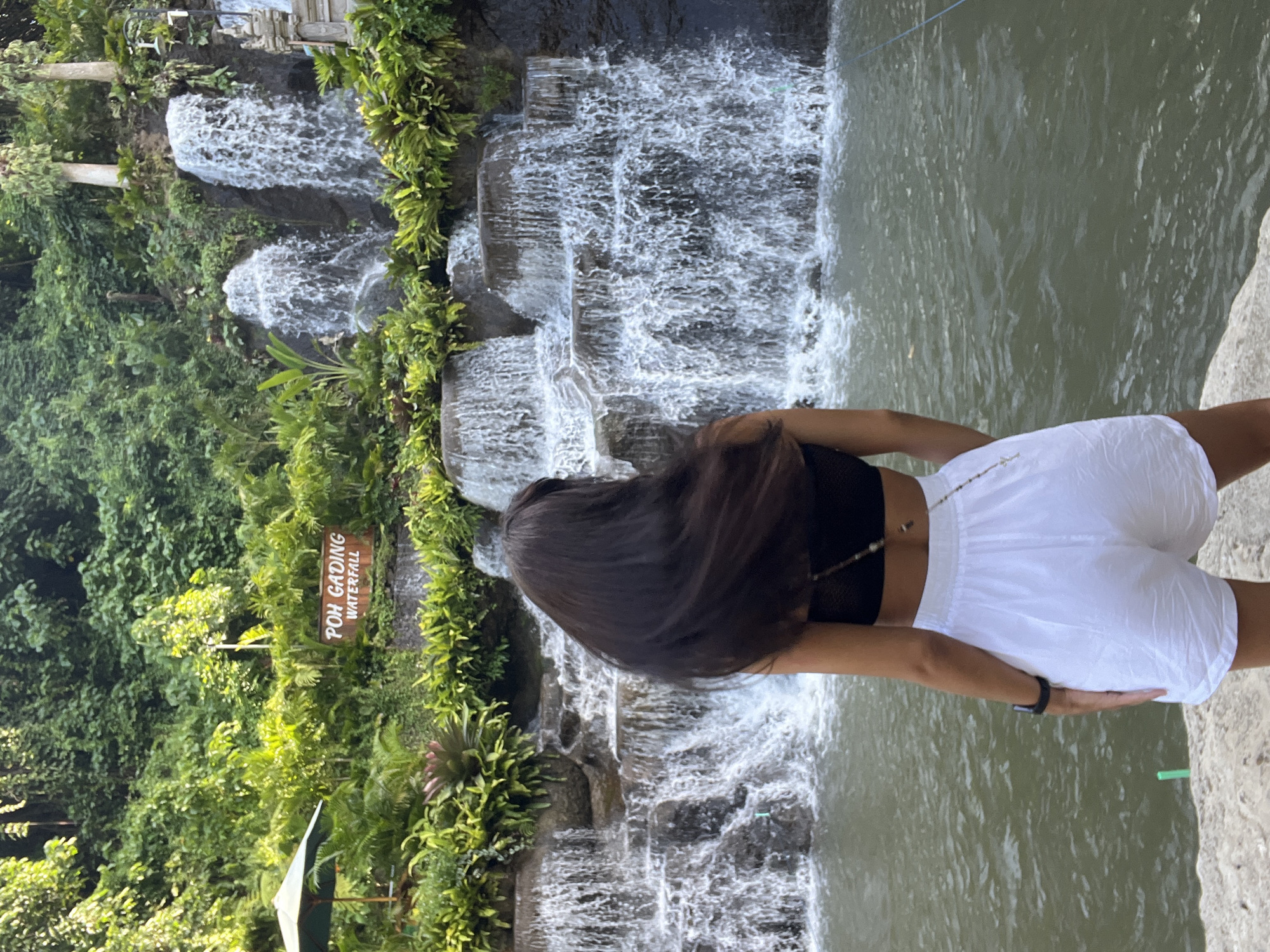 The image depicts a partially visible person with long, dark hair, wearing a black top and white shorts, standing near a waterfall in Sukawati, Indonesia. The waterfall consists of multiple tiers, surrounded by lush green vegetation. A sign reading "Poh Gading Waterfall" is visible amongst the greenery. The water flows into a pool, and part of a stone bank can be seen on the right side of the frame.
