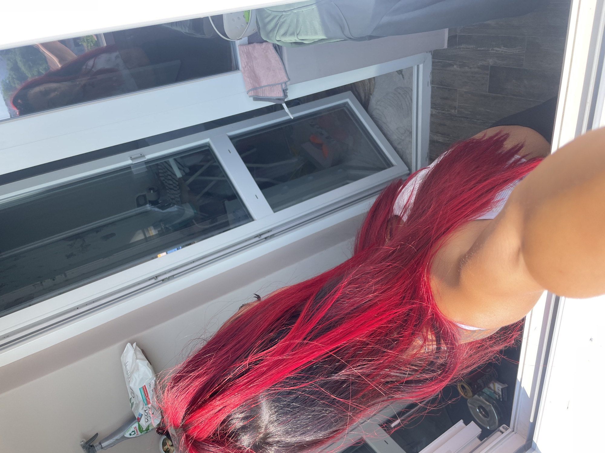 The image shows a person with long, bright red hair, viewed from behind and slightly from the side. The person is wearing a white top. They appear to be standing on a balcony or near an open window. The background shows an outdoor scene with trees visible through a reflective window. There are also some objects on a shelf or ledge, including a white plastic bag with some text that is difficult to read, and a metal object. The lighting suggests it is daytime, likely sunny. The scene is outdoors, possibly in a residential area given the window and balcony. The overall impression is casual and relaxed.
