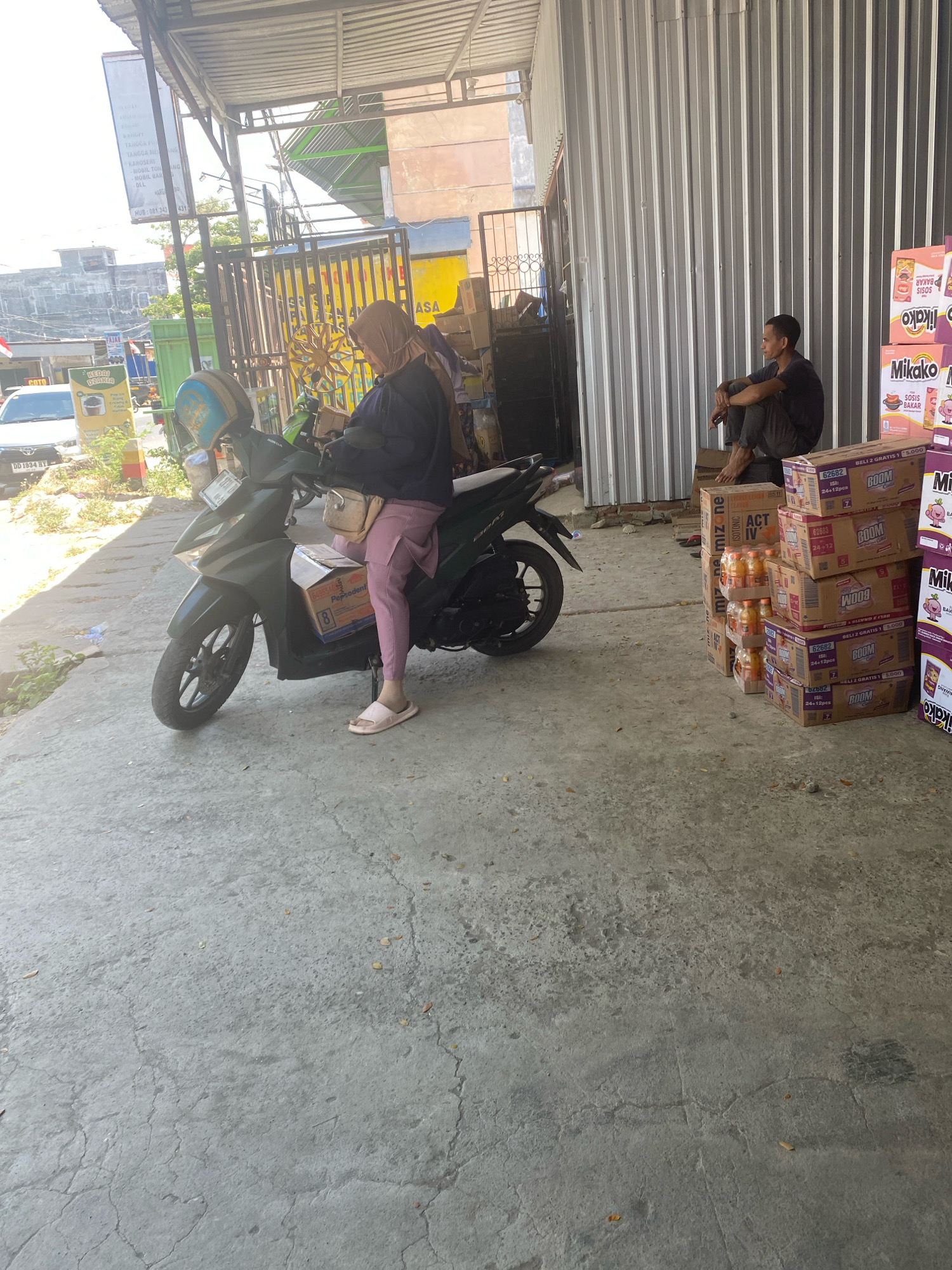 In Kalumeme, Indonesia, a woman wearing a tan hijab and pink pants is seated on a dark-colored motorcycle parked on a cracked concrete surface. She is holding a small box with the text "Pepso dent" visible on it. The motorcycle has a light blue and yellow helmet resting on its handlebars. To the right, a man in a dark t-shirt and pants sits on a stack of boxes near a corrugated metal building. The boxes are stacked high and some are labeled with "Mikako" and "BOOM." The scene appears to be an outdoor area in front of a shop or warehouse, with a sign visible in the background that has Indonesian text and what looks like a phone number. The lighting suggests it is daytime.