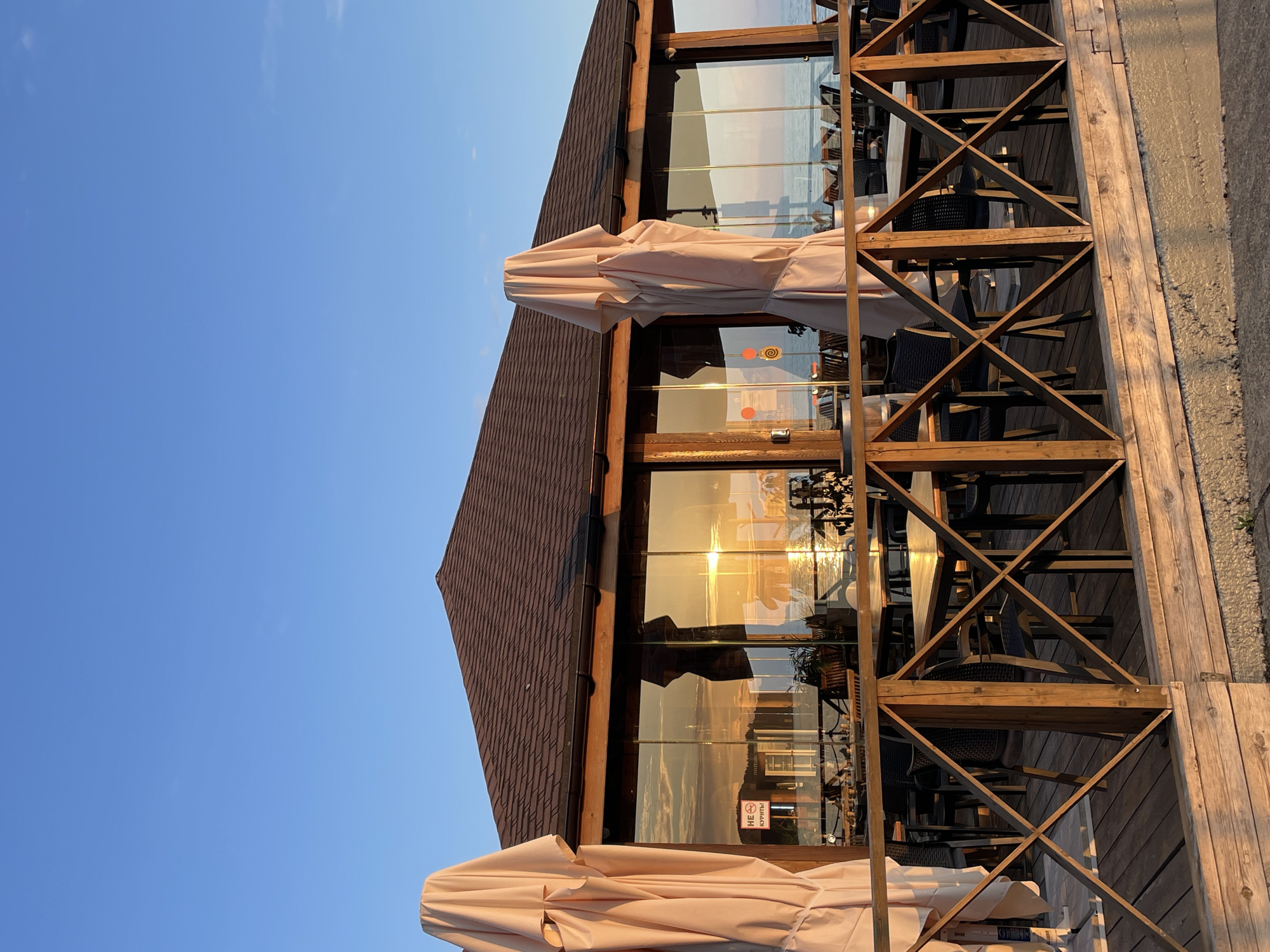 The image captures a restaurant with a wooden facade and large glass windows reflecting the sunset over a body of water. Several tables and chairs are visible through the windows, set up for dining. The exterior features a wooden deck with a railing system composed of crisscrossed beams. Two large, beige umbrellas are folded and tied, suggesting they are not in use. The sky is a clear, bright blue, indicating good weather. The reflection in the windows reveals the warm hues of the setting sun, casting a golden glow on the scene. On one of the windows, a sign with a red circle and a diagonal line through a cigarette, along with text that appears to be in Cyrillic, indicates a no-smoking policy. The overall atmosphere is serene, with the soft light of dusk creating a peaceful ambiance. The location is contextually identified as Sochi, Russia.
