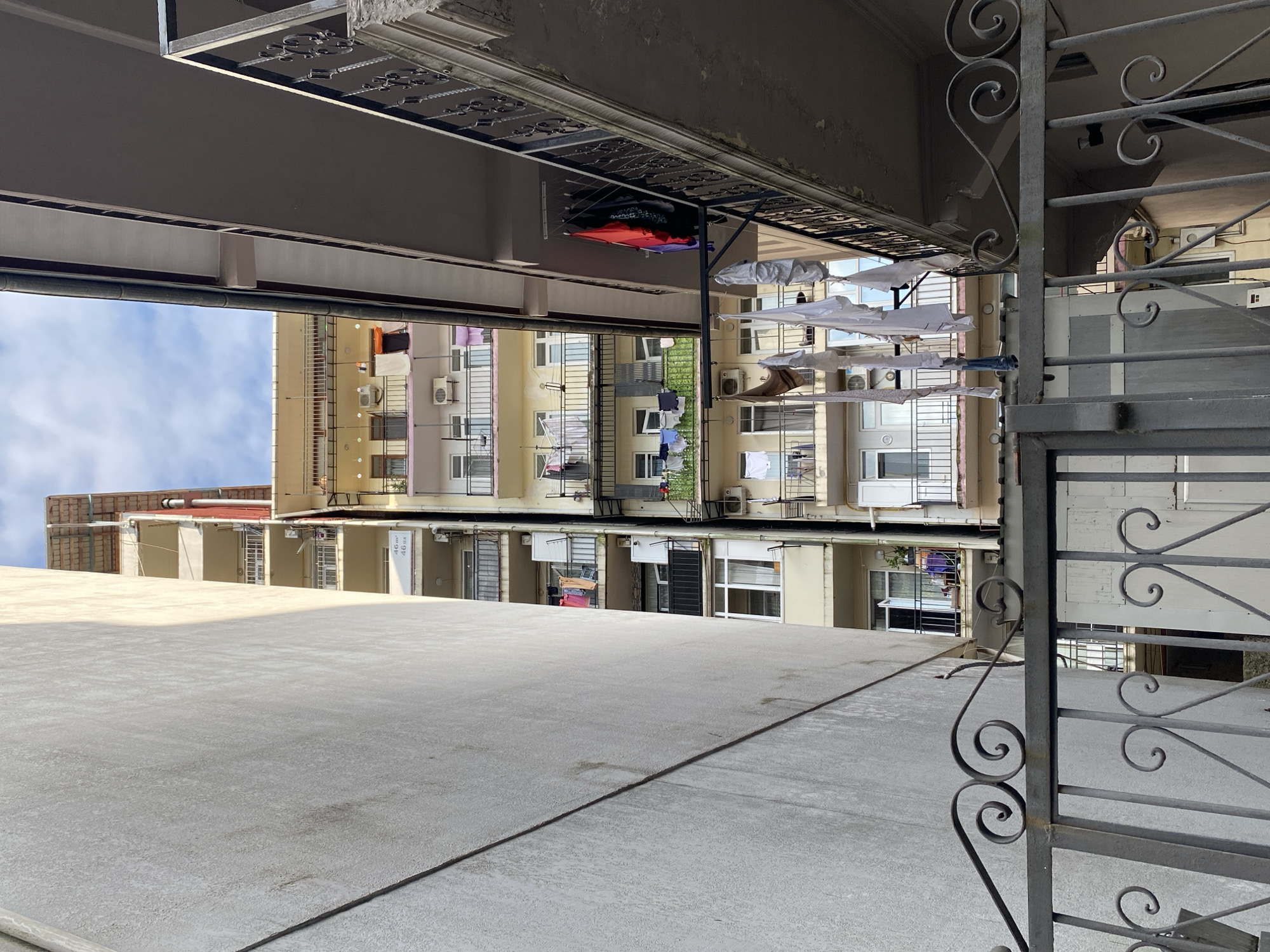 This image shows a view from behind a decorative metal gate looking up at apartment buildings in Batumi, Georgia. On the left, a plain, light-colored wall takes up a significant portion of the frame, casting a shadow. To the right of this wall, a tall, narrow apartment building is visible, characterized by multiple floors with balconies. Laundry is hanging from lines on several balconies, with various items of clothing visible. Air conditioning units are mounted on the building's facade.
To the far right of the image, another apartment building is partially visible, featuring a dark-colored balcony with a wrought-iron railing. More laundry is hanging from a line on this balcony. The sky above is partly cloudy. The overall scene depicts a dense urban environment with a common practice of drying laundry outdoors.