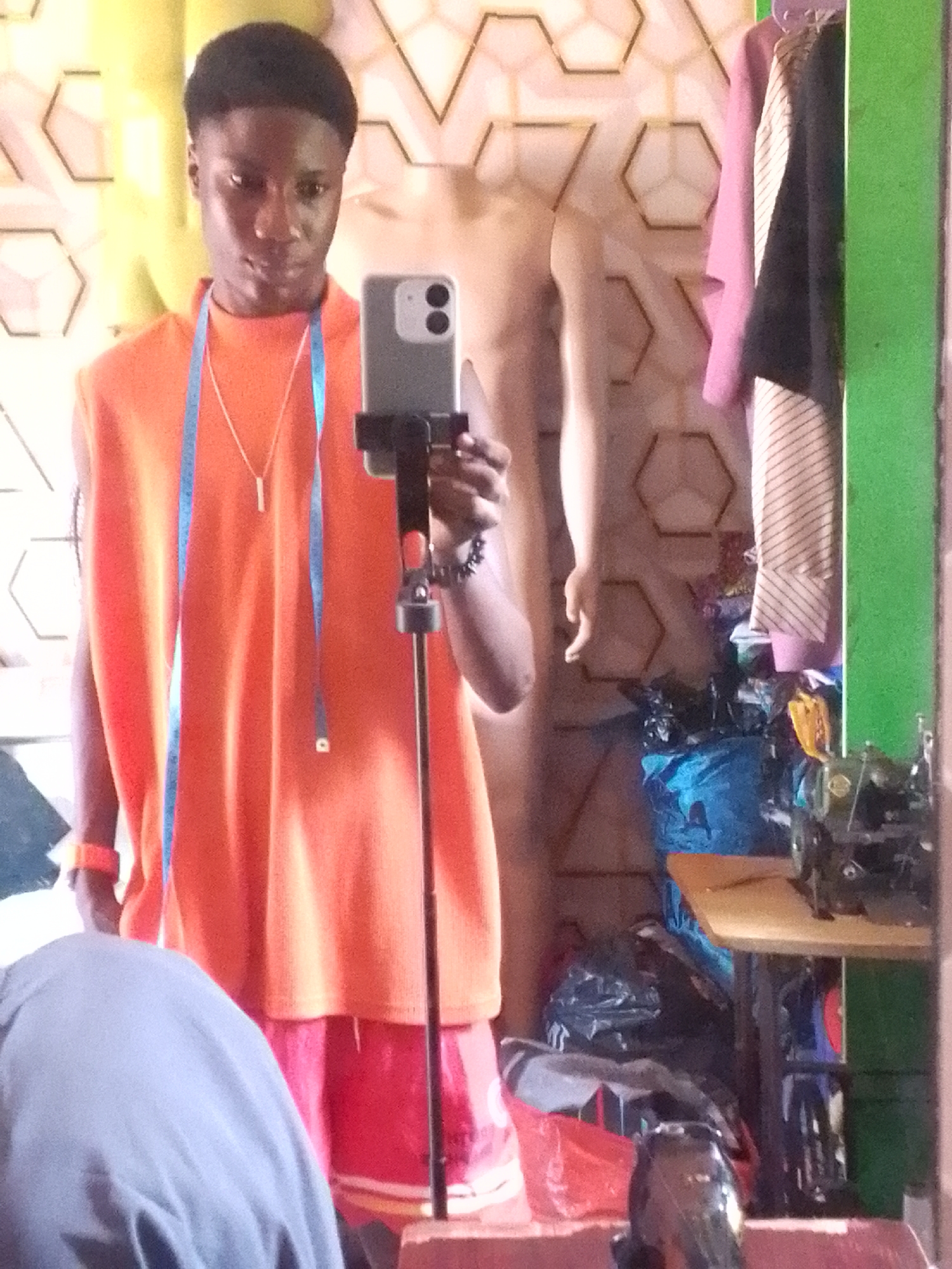 The image depicts a person standing in front of a mirror, seemingly taking a selfie. The individual is wearing an orange sleeveless shirt, red shorts, and a measuring tape is draped around their neck. They are holding a smartphone mounted on a stand. In the background, a mannequin is visible behind the person, along with clothing hanging on a rack, and a cluttered work area with a sewing machine. The mirror's frame is green. The specific location in the city of Unknown, Unknown is unclear, but the setting appears to be a tailor's workshop.