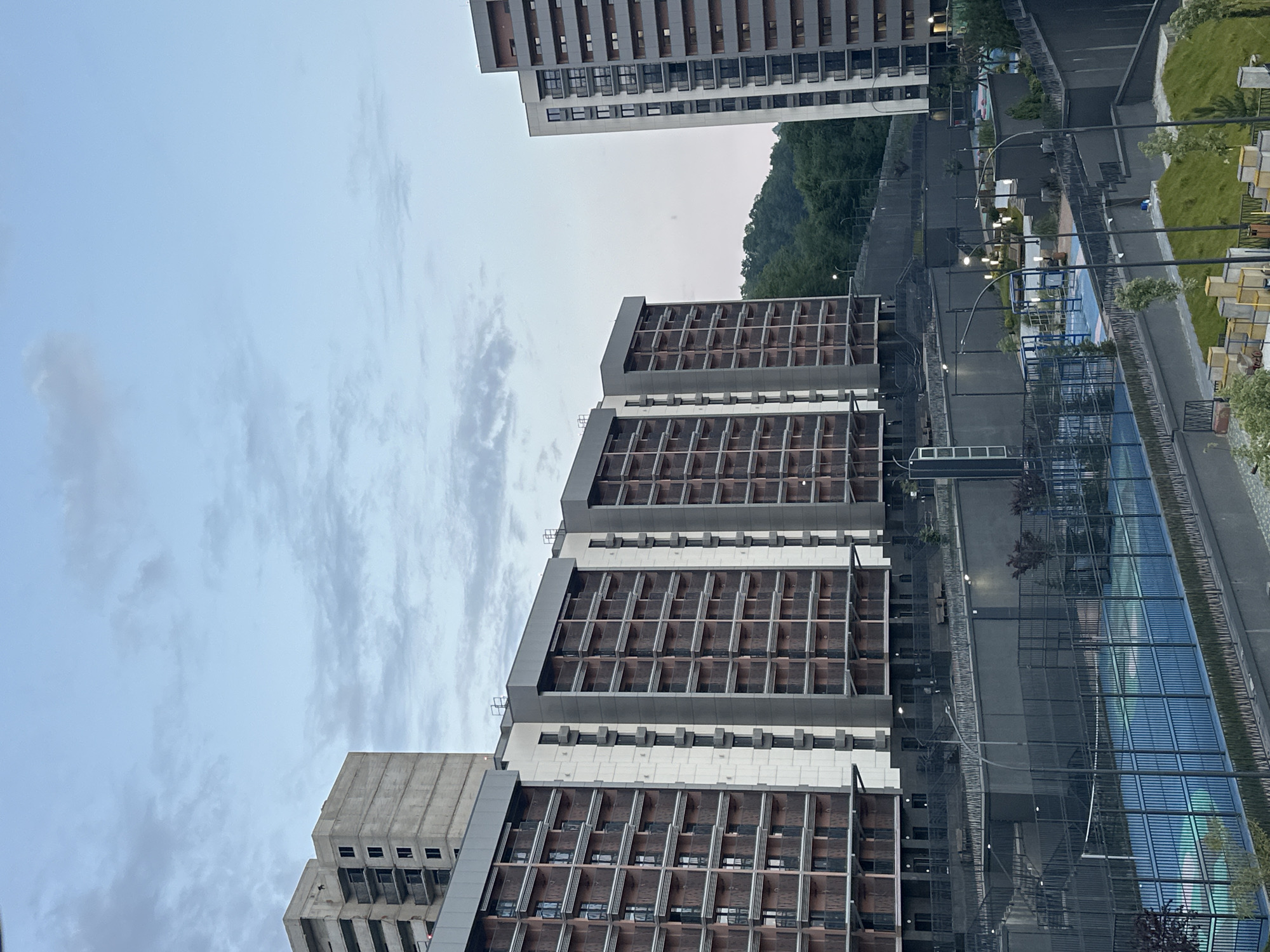 The image captures a residential complex in Razdolnoye, Russia, under a pale, cloudy sky. Multiple tall, modern apartment buildings dominate the scene, some appearing to be under construction with exposed concrete and scaffolding. The buildings feature numerous balconies and large windows, with dark-colored railings.
In the foreground, a fenced area contains a blue sports court, possibly for tennis or padel, with a net visible. The court is surrounded by dark fencing and low walls. Beyond the court, several outdoor lights are visible on poles, illuminating pathways and seating areas. The landscaping includes manicured grass, trees, and concrete barriers. In the background, a dense green forest covers a hillside, contrasting with the urban architecture. The overall impression is one of a developing urban environment integrated with natural surroundings. No people are visible in the image.