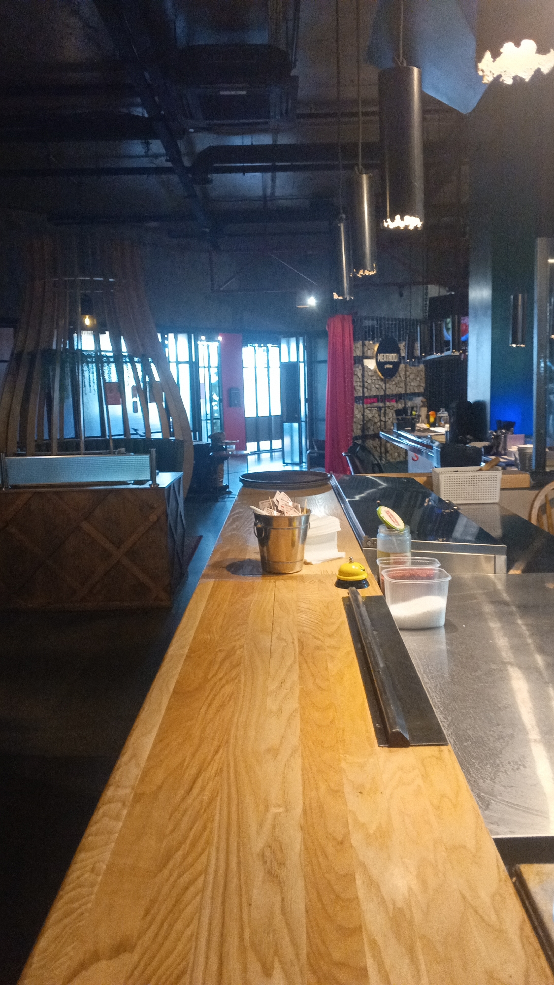 The image depicts the interior of a restaurant or bar, specifically showing a wooden bar top in the foreground. Behind the bar, there are stainless steel surfaces, containers of white granular material, and a small yellow bell. Further back, a large window or set of doors reveals an outdoor area. The ceiling features exposed industrial elements like pipes and light fixtures. A sign reading "MEATHOO" is visible on a patterned wall in the background. The overall ambiance suggests a modern, possibly casual dining establishment. The image was taken in the city of Mytishchi, Russia.