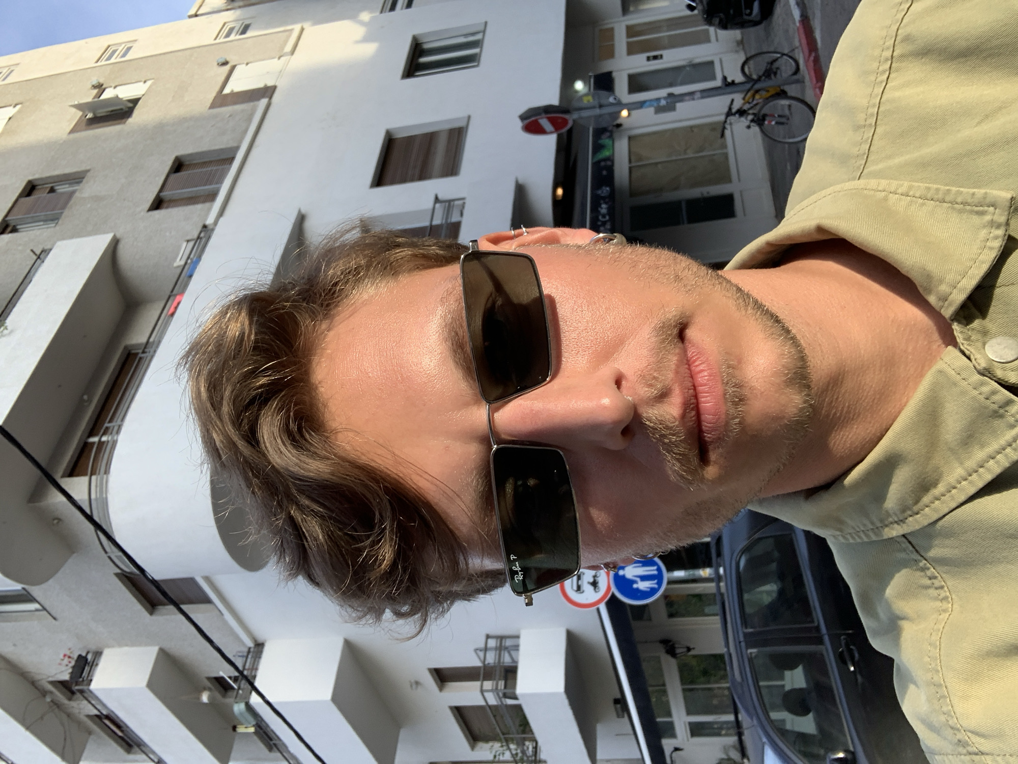 A medium close-up shot depicts a man with light brown hair, wearing sunglasses, and a light green shirt. The man's face is partially obscured by the sunglasses, but his stubble and fair skin are visible. He has multiple piercings in his ear. The background reveals a city scene with white buildings, likely within an urban setting. Road signs with pictograms in blue and white can be seen. A bicycle is also present in the background. Based on the architectural style and signage, the location appears to be in the city of Tel-Aviv, Israel.