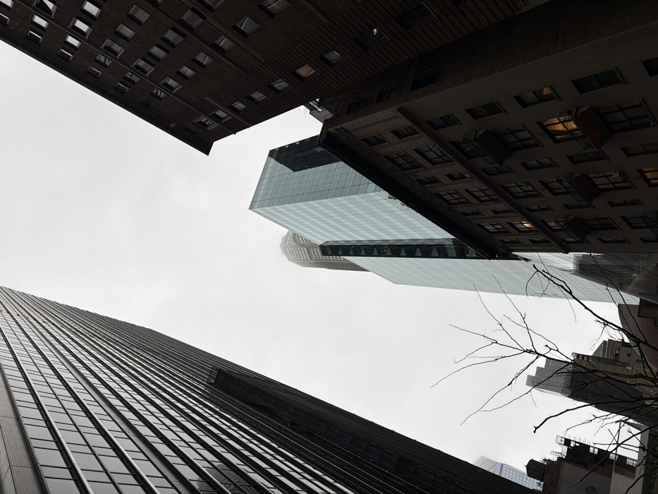 The image presents an upward-angled view of a street canyon in Midtown Manhattan, New York City, United States of America. The sky is uniformly light grey and overcast.
On the left, a tall, dark skyscraper, identified as 1285 Avenue of the Americas, features a facade of reflective dark glass and dark metal panels arranged in a vertical grid pattern, extending diagonally from the lower left upwards.
In the mid-ground, a modern skyscraper with a light blue-green glass facade, identified as 1211 Avenue of the Americas, rises vertically. Behind it, the upper section of another very tall, dark-toned skyscraper, likely 1221 Avenue of the Americas, is partially obscured by atmospheric haze.
On the far right, an older, multi-story building constructed of dark brown brick is visible, featuring numerous rectangular windows. Several windows on this building, and on a lighter-toned, multi-story building partially visible below it, display interior illumination.
In the lower center foreground, a bare, deciduous tree branch with multiple fine twigs is silhouetted. No individuals, vehicles, or discernible actions are present.