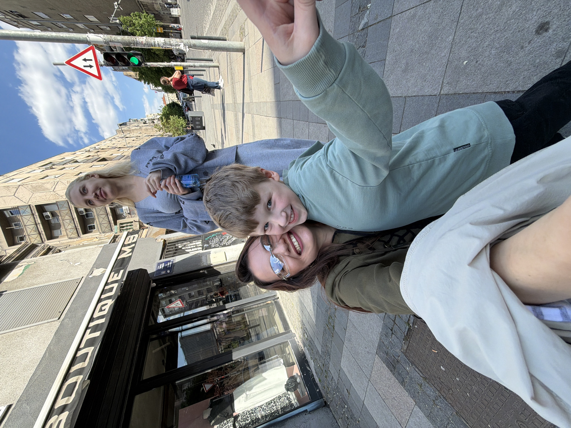 This media file captures a cheerful selfie taken by a group of three individuals on an urban street, presented in a sideways orientation. The scene is bright, suggesting daytime with clear, sunny weather, indicated by a blue sky with scattered white clouds.
In the immediate foreground, a woman with dark hair and sunglasses smiles widely at the camera. Next to her, a young boy with light brown hair and blue eyes grins, leaning his head against hers. He wears a light green long-sleeved top, with a small black tag that reads "crocKid ®" visible on the sleeve. Behind them, a young woman with blonde hair, dressed in a grey hoodie, also smiles towards the camera, holding a small blue bottle in her hands.
The background reveals a bustling city street. Paved sidewalks run along the side of multi-story buildings with classic urban architecture, some showing visible air conditioning units. A prominent feature in the background is a traffic pole with a triangular road sign displaying two arrows (one up, one down, typically indicating "Priority to oncoming traffic" or "Give Way"), situated next to a traffic light that is currently green. Further back, another person in a red shirt and blue jeans can be seen walking. On the left side of the frame, an awning or sign for a shop is visible, with the text "BOUTIQUE MS" clearly readable. The overall atmosphere conveyed by the smiling faces is one of happiness and enjoyment.