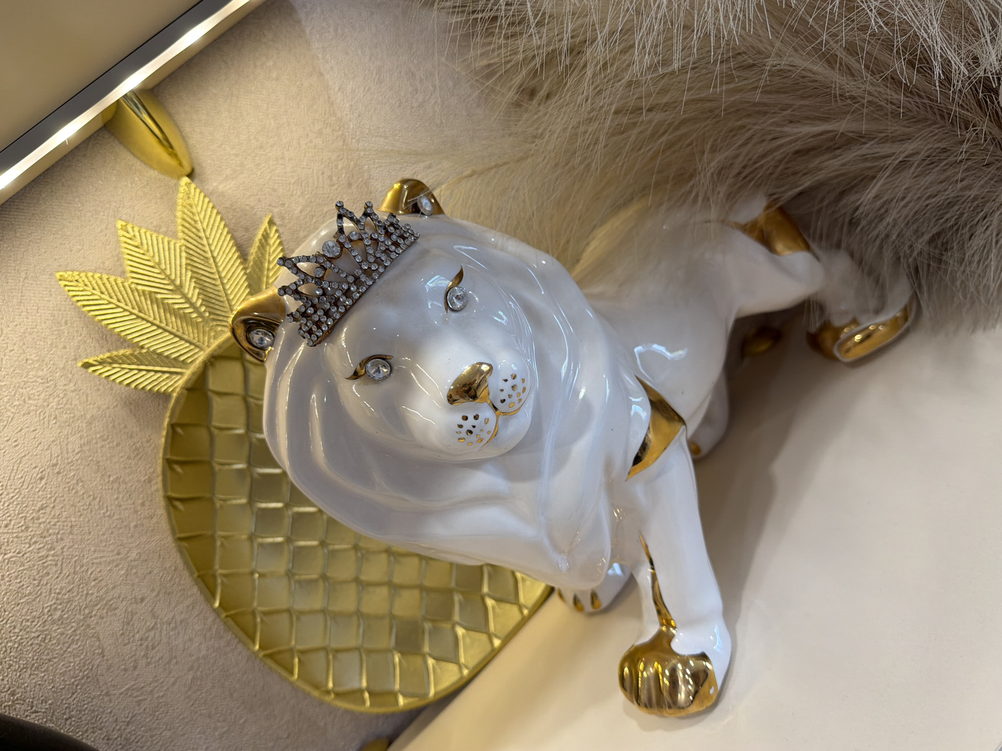 A close-up, high-angle shot captures a white ceramic lion figurine adorned with gold accents and a sparkling crown. The lion, facing slightly towards the right, has a polished, glossy finish. Its paws, ear tips, and details around its eyes and nose are painted gold. Two large clear stones serve as its eyes, while its nose and forehead feature additional small clear stones, culminating in a prominent diamond-studded miniature crown perched on its head.
Behind the lion's left shoulder, a golden, pineapple-shaped decorative element with a textured, grid-like pattern is visible, serving as a backdrop or tray. To the right and behind the lion, a fluffy, light-colored material, resembling faux fur, provides a soft, textural contrast. The lion appears to be resting on a light-colored, possibly white, surface.
In the upper left corner, a modern linear light fixture, possibly an LED strip, is mounted on a textured, light-beige wall, which is also visible in the background behind the pineapple. The lighting suggests an indoor setting, likely under artificial illumination. There is no discernible activity or visible text in the image. The arrangement gives a sense of decorative luxury, possibly from a retail display or home decor in Dzerzhinsk, Russia.