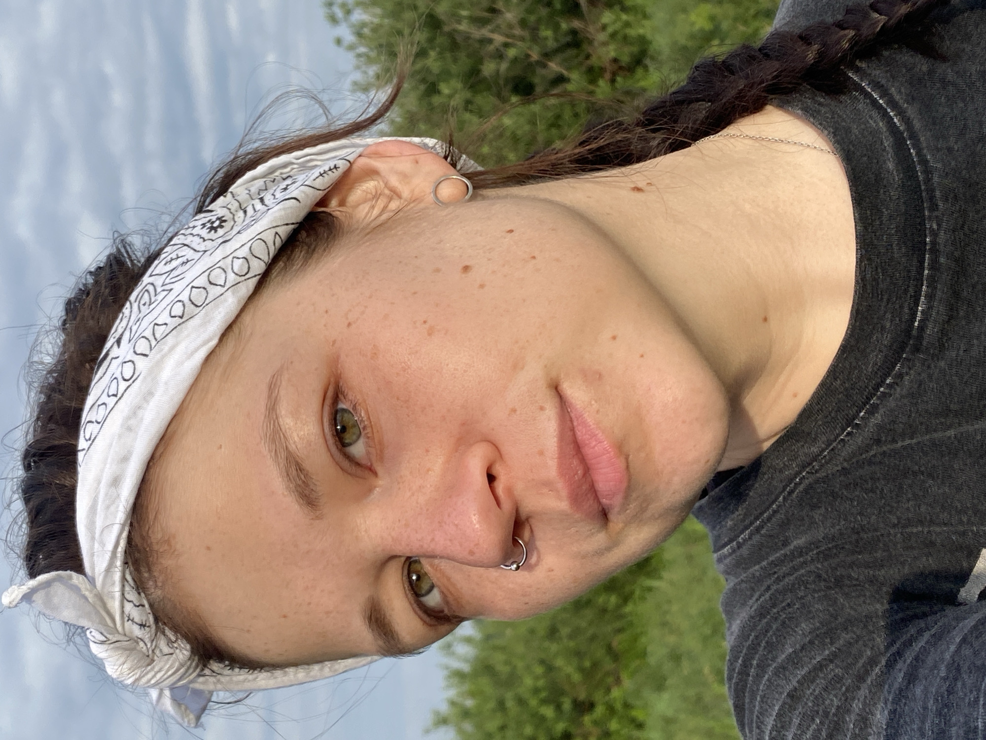 A close-up, eye-level, outdoor selfie shows a young woman from the chest up. She is looking at the camera with a neutral expression. Her hair is dark brown and pulled back with a white bandana patterned with black paisley designs. She has green eyes, freckles across her nose and cheeks, and a septum piercing. She is wearing a dark grey t-shirt. The background is blurred green foliage and a cloudy sky. The lighting suggests it is daytime. The location is not specified in the image.