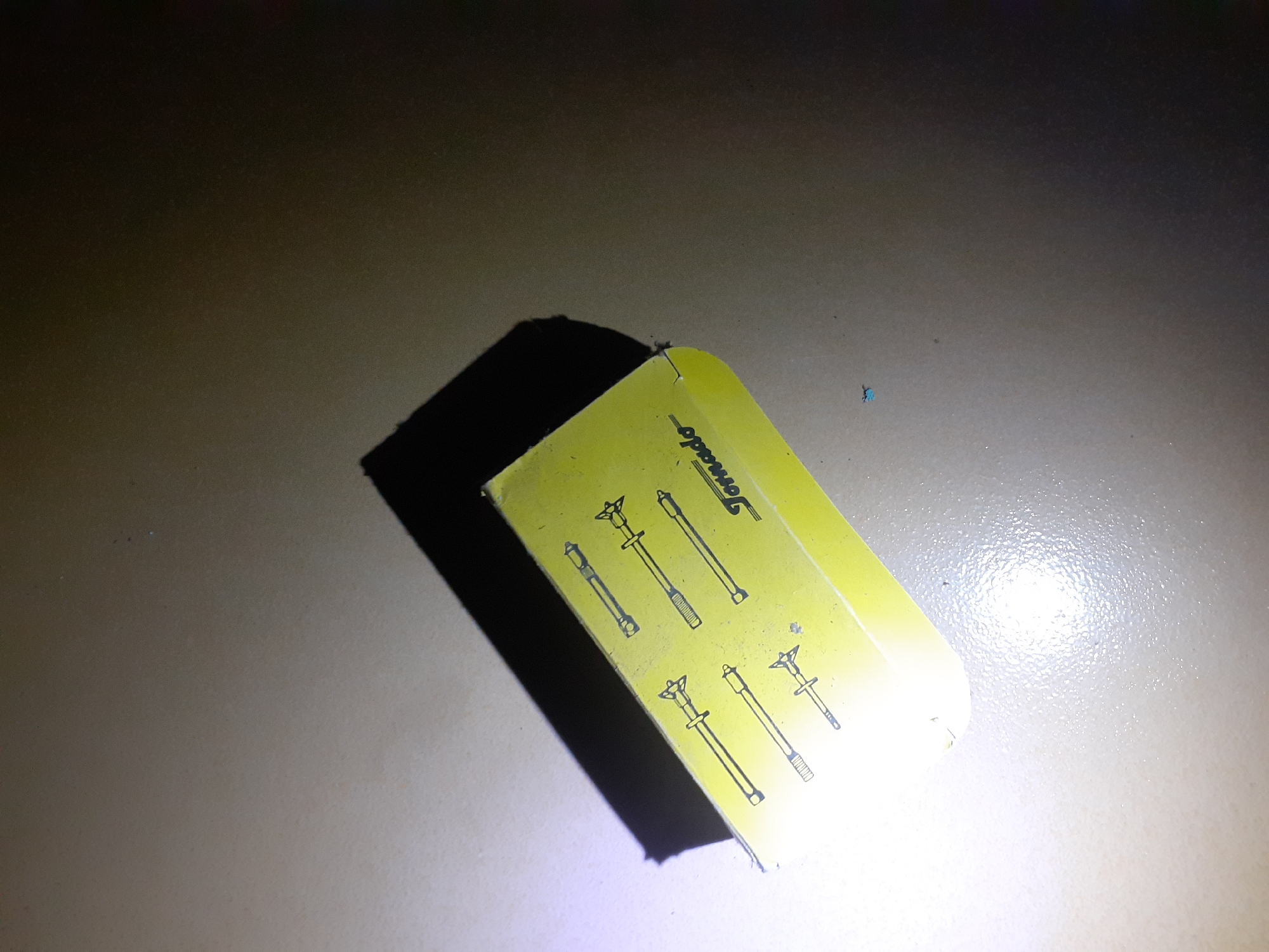 The image shows a yellow box with black line drawings of what appear to be tools or components. The word "Tornado" is printed in black script on the lower right portion of the box. The box is casting a shadow on a textured, beige-colored surface. The lighting is uneven, with a bright light source in the lower left corner causing glare and a dark shadow on the upper right. There are no people visible in the image. The scene appears to be an indoor setting, possibly a table or countertop. The specific location in Jalingo, Nigeria, cannot be determined from the visual information provided.
