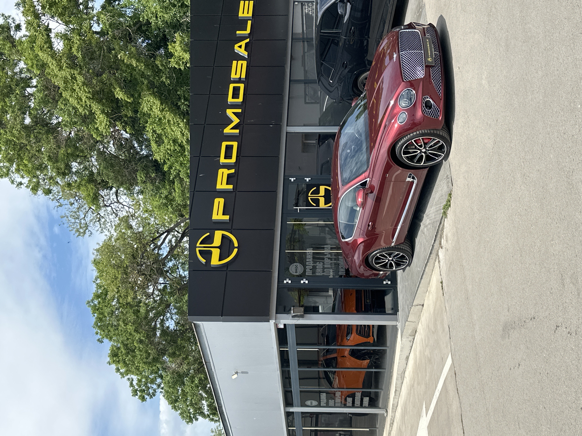 A photograph shows a burgundy Bentley automobile parked in front of a dealership with the name "PROMOSALE" displayed in large yellow letters. The car is positioned at an angle to the building. To the left of the dealership is a tree with green foliage. The sky is visible in the upper left corner, with a mix of blue and white.
The dealership has a modern design, with glass windows showcasing cars inside, including an orange vehicle. The location is in Varna, Bulgaria, as evidenced by the text displayed on the dealership window in Bulgarian, suggesting the business operates there. The image is captured during daylight hours.