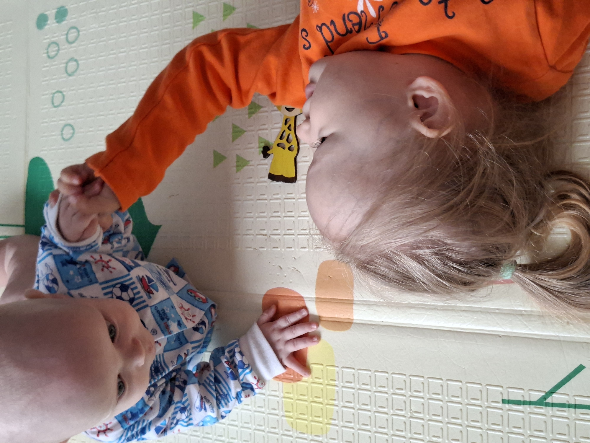 Two young children are lying on a play mat. One child is wearing an orange long-sleeve shirt with a giraffe design and text, while the other is wearing a blue and white patterned long-sleeve onesie. The children are holding hands. The play mat has a variety of shapes and colors. The setting is in an unspecified location in an unknown city.