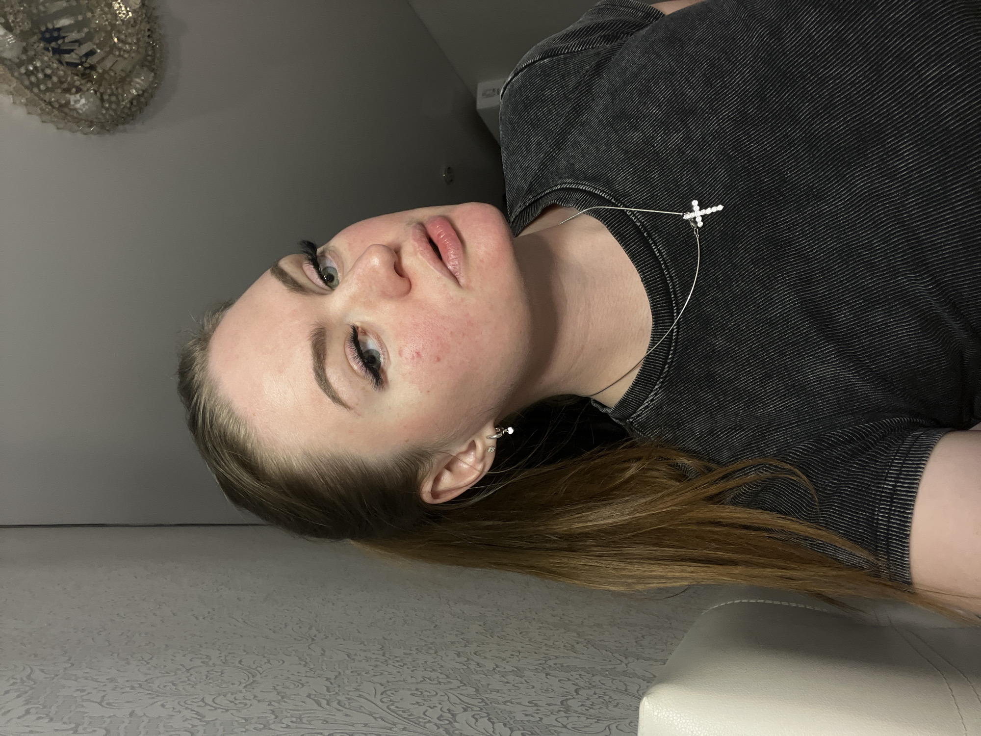 An indoor close-up portrait features a young woman lying horizontally across the frame, her head positioned on the left and facing towards the top, set in Sevastopol, Ukraine. She has light skin with some visible redness on her cheeks and chin. Her light-colored eyes are open, and she has pronounced dark eyelashes, possibly extensions. Her eyebrows are well-defined. Her lips are slightly parted, revealing a natural pink tone. Her medium brown hair is pulled back, extending towards the bottom-right of the frame.
She is wearing a dark, possibly grey or black, ribbed t-shirt or top. A delicate silver necklace with a sparkling cross pendant rests on her chest, and a small silver hoop earring with a dangling charm is visible in her right ear.
The background includes a plain light grey wall. In the upper left corner, a decorative object, possibly made of silver or crystals, is partially visible. The surface directly beneath the woman's head and hair is a light-colored material with a subtle, ornate pattern, suggesting wallpaper or flooring. Part of a light, upholstered piece of furniture, like a chair or sofa, can be seen in the bottom right corner. The lighting appears soft and even, typical of an indoor setting with artificial light. The woman's expression is neutral and composed. There is no visible text in the image.