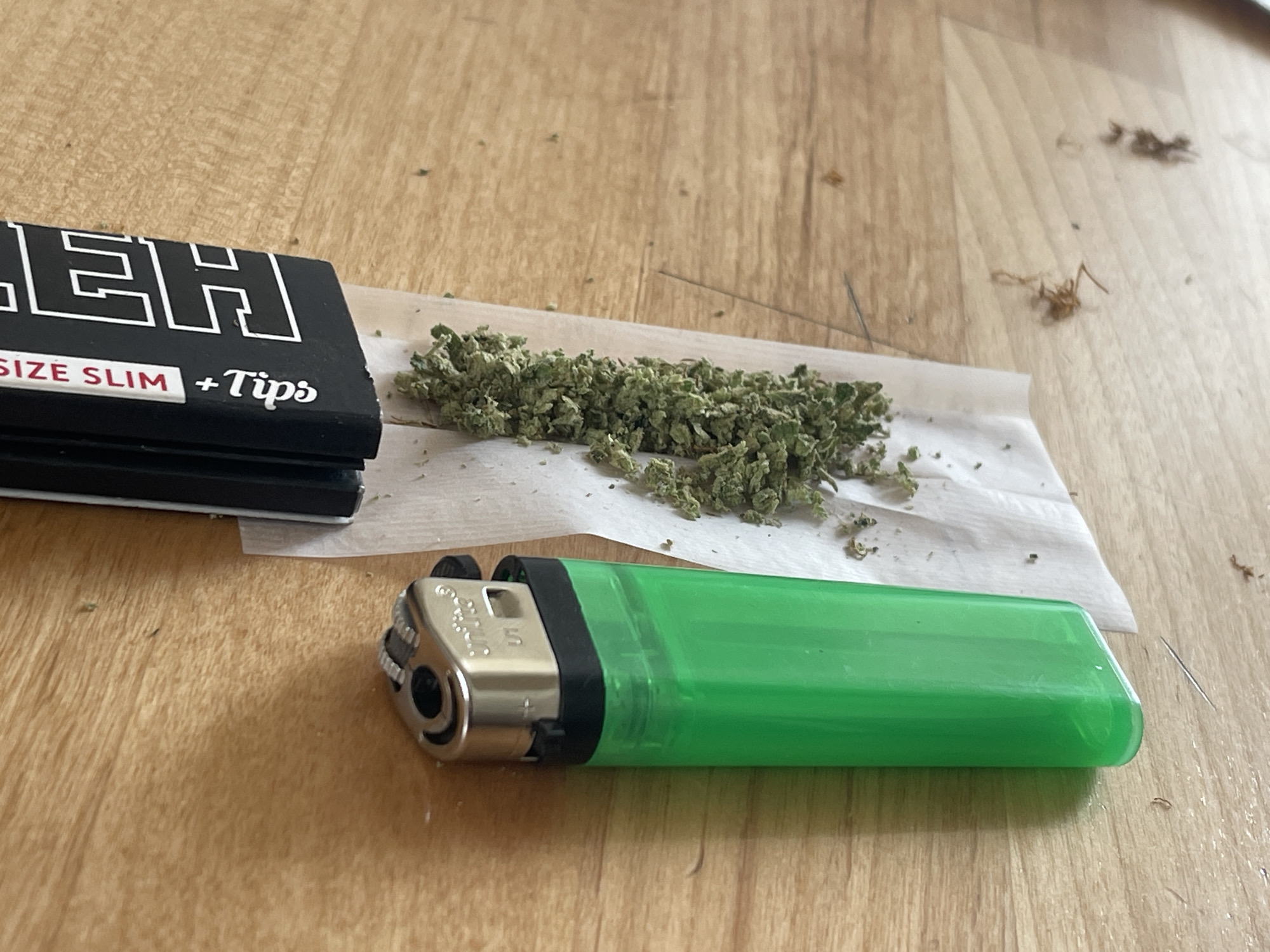 In an indoor setting with a light-colored wooden floor, a pile of marijuana rests on rolling papers. To the left of the marijuana is a black package of rolling papers with white text that reads "SIZE SLIM + Tips". In the foreground, a bright green lighter is positioned horizontally. The scene is lit by overhead lighting, casting soft shadows. The image is a close-up shot, with the camera focused on the objects. No people are visible in the frame.