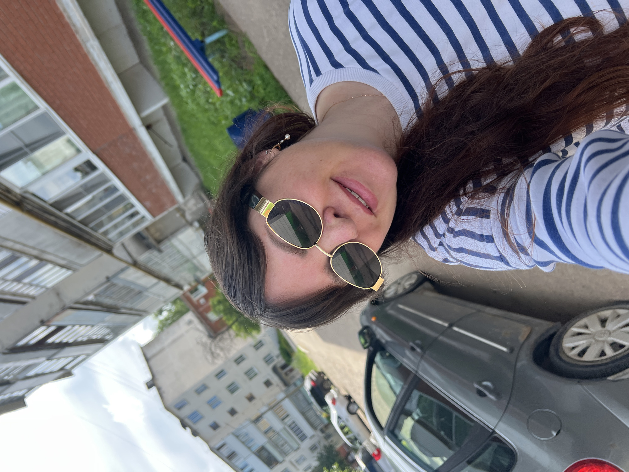 A woman with long, dark brown hair, wearing a white long-sleeved shirt with blue stripes and gold-rimmed sunglasses, is captured in a selfie. She is standing outdoors in what appears to be a residential urban area. In the background, several apartment buildings with visible windows are present. To the right of the woman, a dark grey car is parked on the street. The scene suggests a casual outdoor moment in a city like Noviy, Russia, possibly on a street or in front of a residential complex.