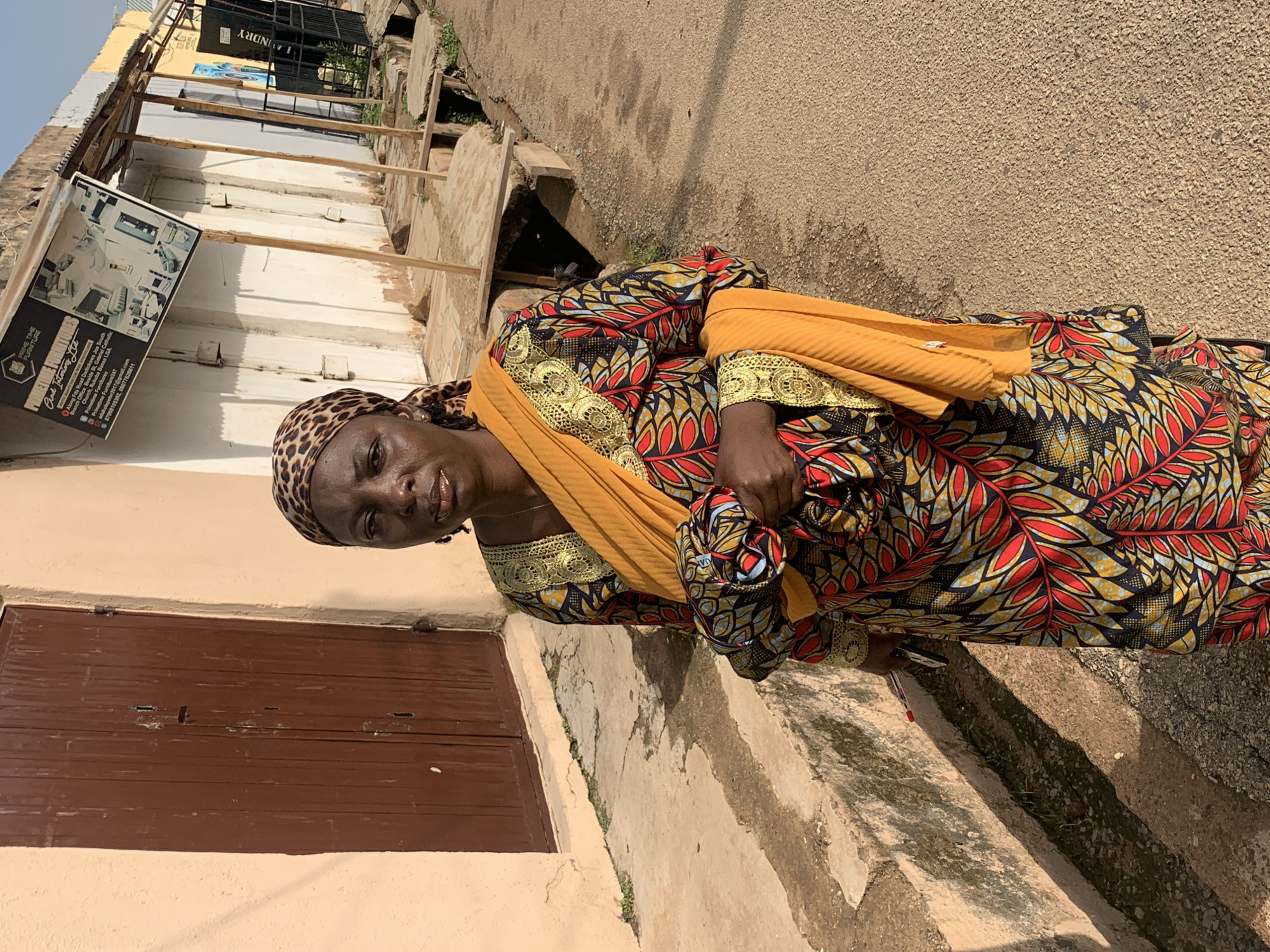 A medium shot shows a Black woman standing on a dirt road in Jos, Nigeria. She is wearing a vibrantly colored dashiki dress with a yellow sash draped over her shoulders and a leopard-print headscarf. Her arms are crossed in front of her, and she is holding a small object, possibly a phone, in her right hand. She is looking directly at the camera with a neutral expression.
The background consists of a building with a brown wooden door on the left and a storefront with a metal awning on the right. A sign for "PRIME TIME FURNITURE" is visible on the storefront. The ground is a dirt road with a drainage ditch running alongside it. The scene appears to be in an urban or semi-urban area. The lighting suggests it is daytime, with shadows cast by the buildings and the woman.