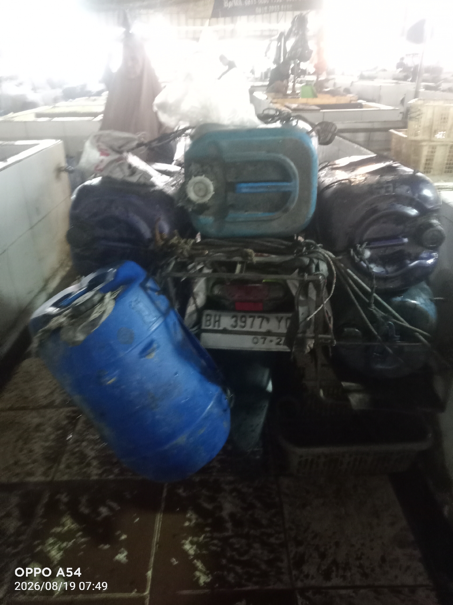 The image depicts a motorcycle parked indoors, likely in a market or storage area, with several large plastic barrels secured to its rear. The barrels are predominantly blue and dark in color. One blue barrel is prominently placed on the left, tilted and appearing damaged with an open top revealing some material inside. Another blue barrel is positioned on top of the motorcycle's rear rack, along with a dark-colored barrel. The motorcycle has a license plate visible with the characters "BH 3977 YO" and "07-24". The environment appears to be a dimly lit indoor space with a tiled floor that shows signs of being wet or dirty. In the background, there are structures that resemble market stalls and some indistinct figures, one wearing a light-colored head covering. The overall impression is one of transport or storage, possibly of liquids or other goods, within a busy or utilitarian setting. The presence of the license plate format suggests the location is in Indonesia, and the "BH" prefix is specific to Jambi province, Indonesia. The timestamp "2026/08/19 07:49" is superimposed on the bottom left of the image, indicating the time and date the photograph was taken.