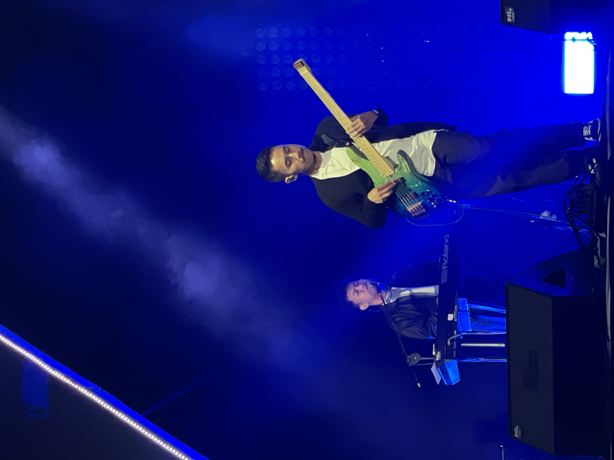 The image captures a musical performance on a dimly lit stage, bathed in blue light. A man stands playing a multi-colored bass guitar. He is wearing a white shirt and a black jacket with black pants. To the lower-right of the guitarist, another man sits at a keyboard, also dressed in a jacket and shirt. A Roland keyboard stand is visible, as is a large black speaker. A white light fixture is visible in the background. The location, Salembaran, Indonesia, is not verifiable from the image alone.