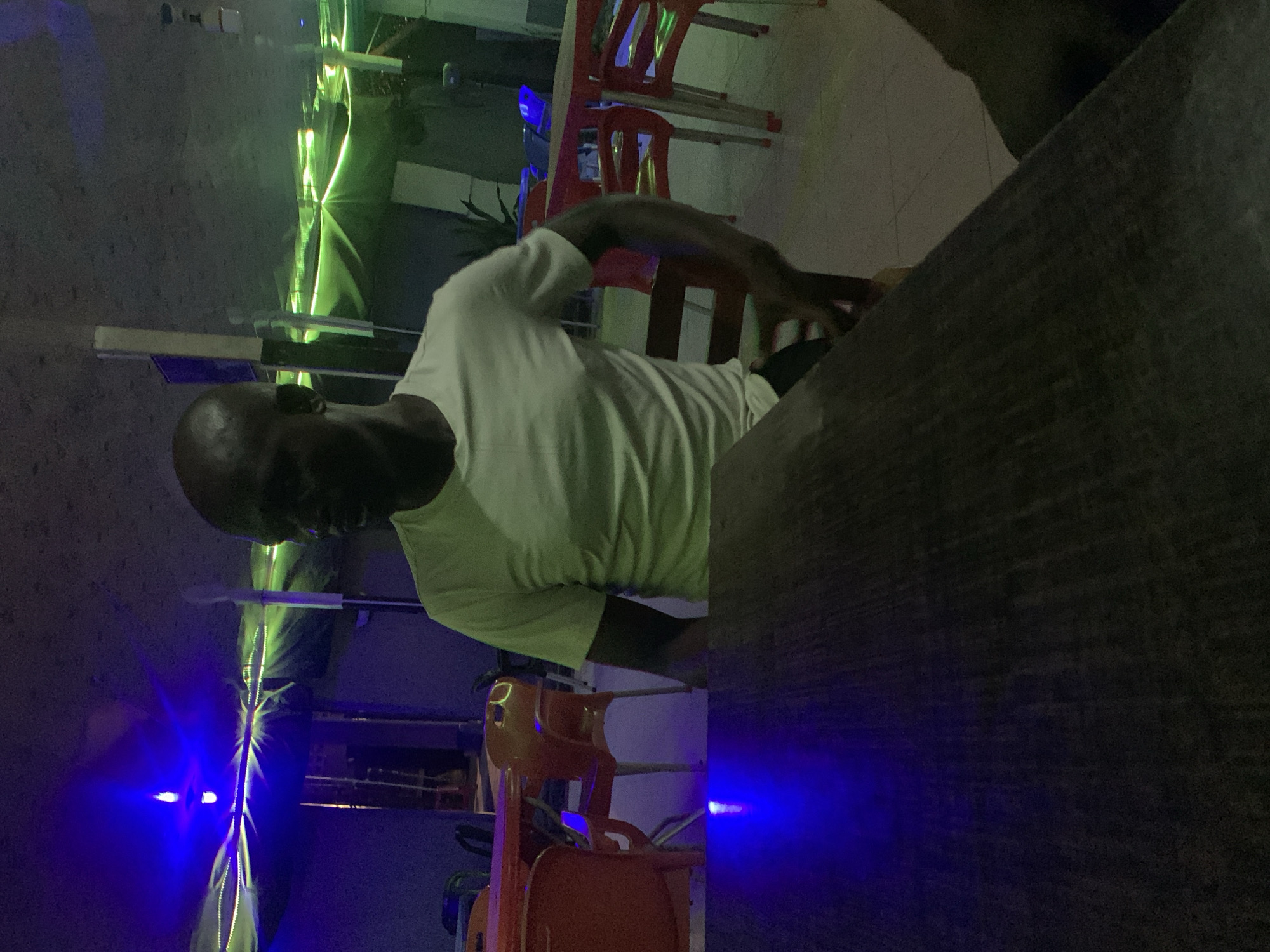 The image, taken in Akwanga, Nigeria, captures a dimly lit indoor or covered outdoor setting at night. The central subject is a dark-skinned man with a bald head, viewed from a slightly elevated angle. He is wearing a plain white short-sleeved t-shirt and is seated at a large, dark table that occupies much of the right foreground. His head is turned downwards or to his left, suggesting he is focused on something on the table or in front of him, though his exact activity is not discernible.
The background is defined by prominent artificial lighting. Vibrant green linear lights crisscross across the mid-ground, creating bright streaks and reflections on unseen surfaces, possibly walls or architectural features. A distinct blue light source glows intensely in the lower left, casting a blue hue onto a dark, textured wall. Additional blue light elements are visible in the distance. Scattered throughout the background are several red and orange plastic chairs, some stacked, implying a casual public venue like a bar, restaurant, or lounge. The overall ambiance is dark and atmospheric, characterized by the interplay of neon light colors against the deep shadows. No visible text is present in the image.