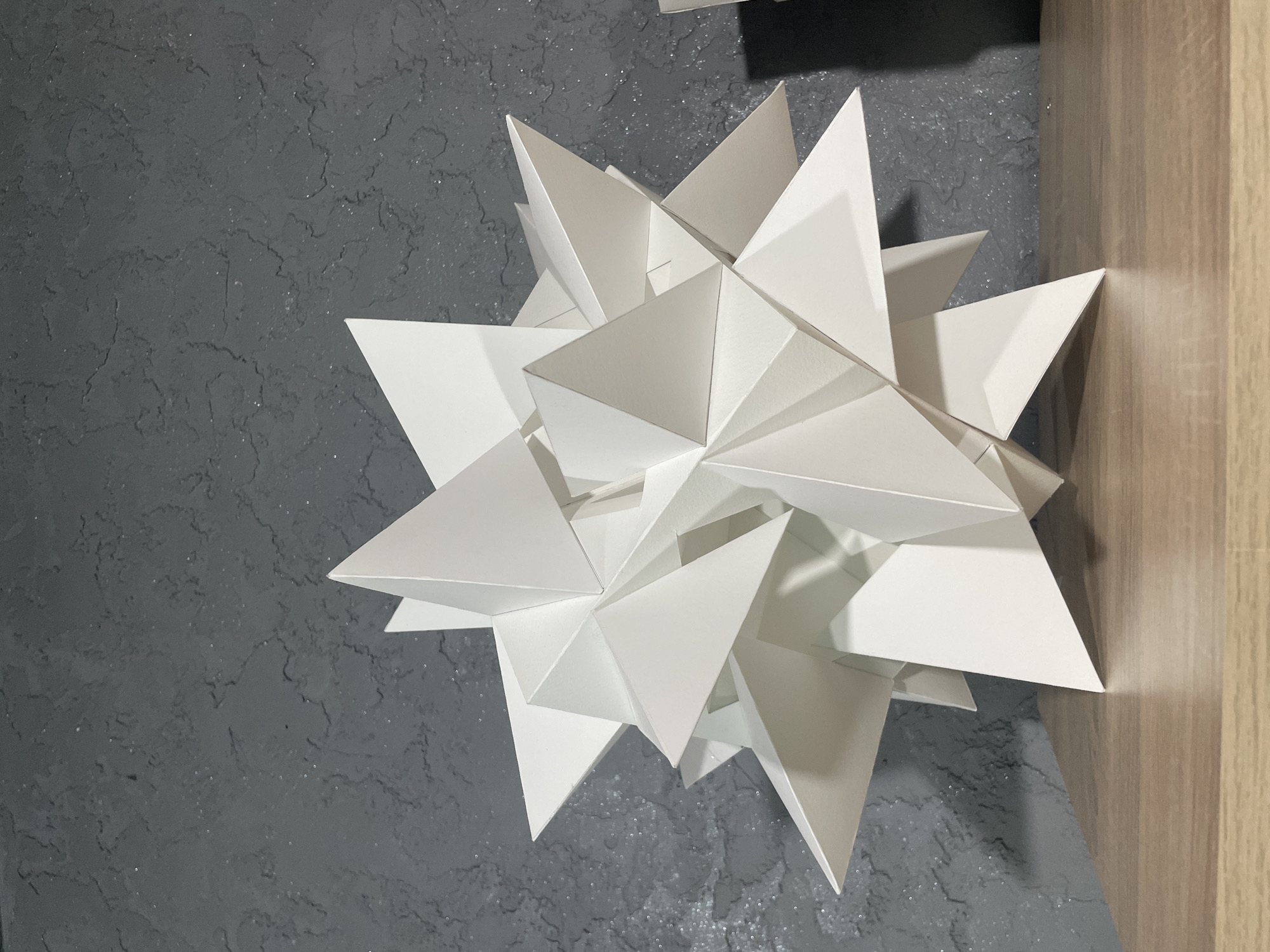 The image depicts a white, geometric paper sculpture resembling a starburst or complex polyhedron. The sculpture is composed of numerous triangular or pyramidal forms joined together, creating a spiky, three-dimensional object. The sculpture is placed against a textured, dark grey wall with an uneven, almost stucco-like surface. To the right of the sculpture, there is a light brown or beige surface, possibly a wooden panel or the edge of a piece of furniture. There is no indication that this image was taken in the city of Sofia, Bulgaria. There are no people visible in the image. The scene is well-lit, highlighting the textures of the wall and the paper sculpture.