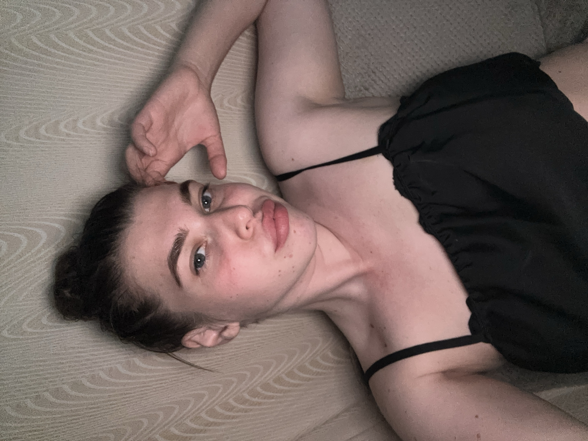 A woman with fair skin and blue eyes is lying on a light beige, patterned surface. She has dark hair pulled back in a bun. She is wearing a black top with thin straps. Her right hand is near her forehead. The location is unknown and there is no indication that this image was taken in Мещёры, Russia.