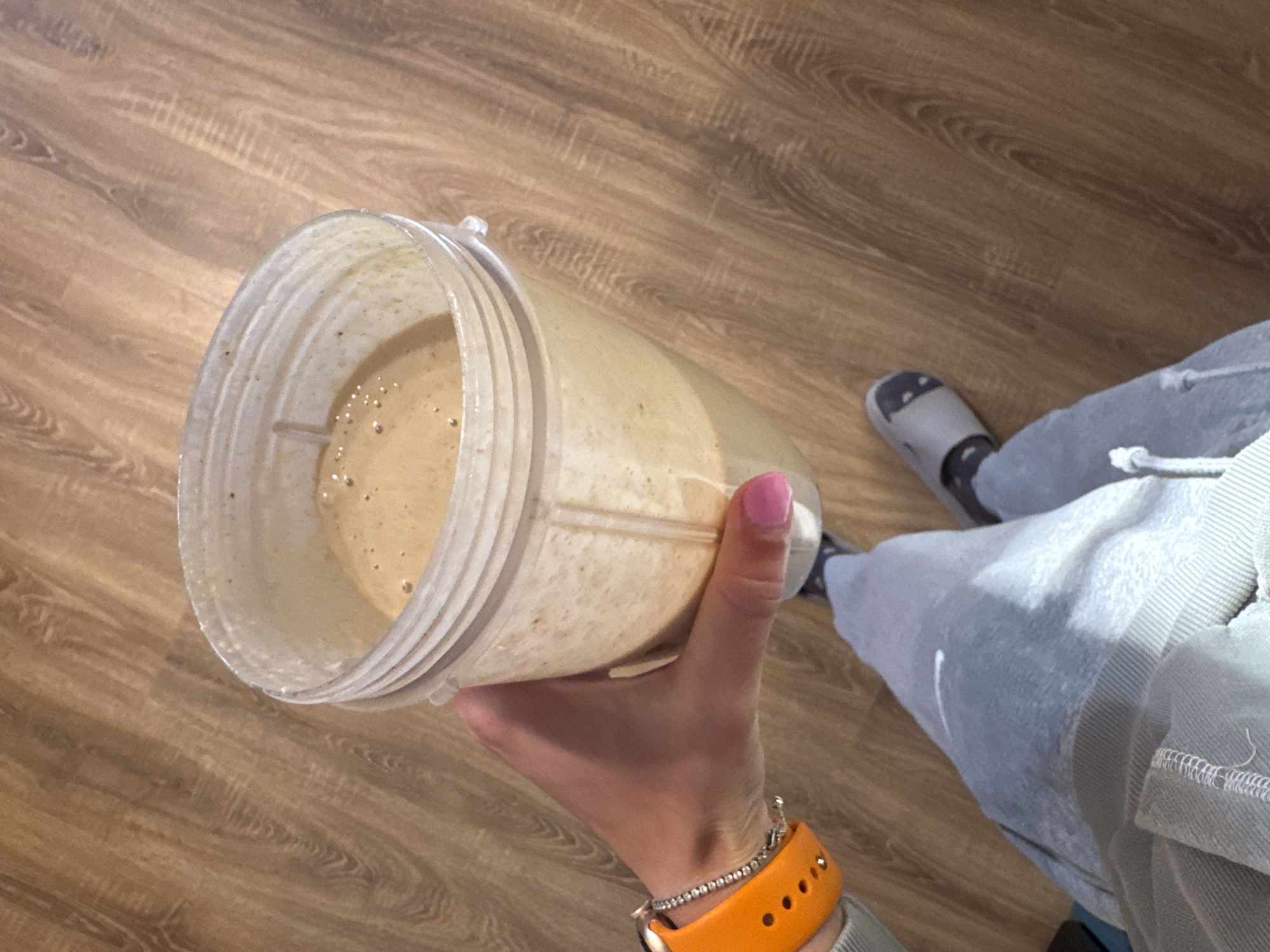 The image depicts a first-person perspective of a person holding a transparent plastic blender cup filled with a light brown liquid, likely a smoothie. The person is wearing light gray sweatpants and a gray slide sandal on their right foot. They have an orange smartwatch band and a silver bracelet on their wrist. The background consists of a wood-look laminate floor. The location is unknown and cannot be determined as Sofia, Bulgaria, without additional information.