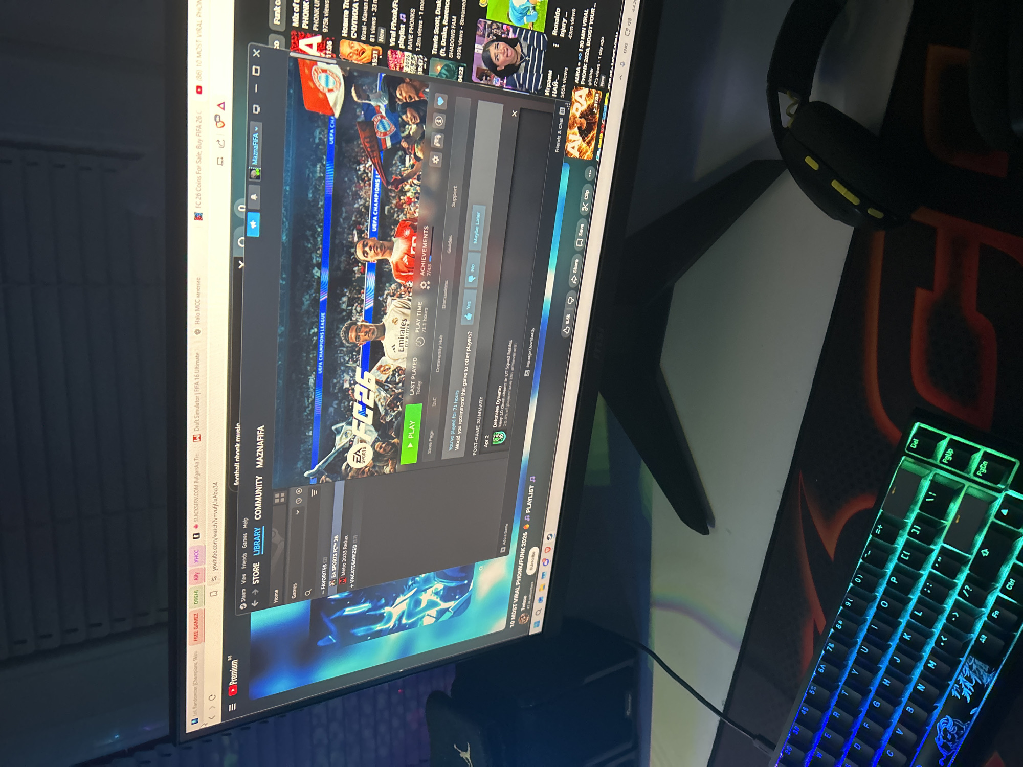 A monitor displays a Steam window with the game "EA SPORTS FC™ 26" open, suggesting the user has been playing it for 71.1 hours. The Steam interface shows tabs for Store, Library, and Community. Multiple YouTube tabs are open in the background, including one titled "10 MOST VIRAL PHONK/FUNK 2026." A black gaming keyboard with illuminated keys and a gaming headset are visible on the desk to the right of the monitor. The URL "SLACKSERV.COM Bulgarska Tre" indicates a Bulgarian context. Given the reference to "Bulgarska Tre" and the Bulgarian words visible in the YouTube video titles, this is likely located in Sofia, Bulgaria.