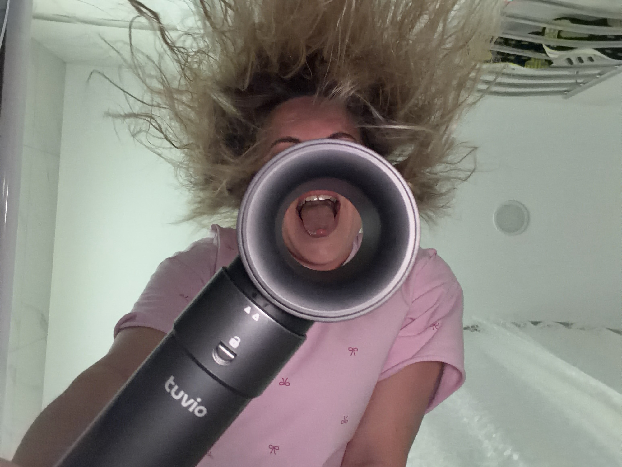 An inverted, upward-angle photograph captures an individual, likely female, with light brown, frizzy hair, holding a dark grey 'tuvio' brand hairdryer. The hairdryer's concentrator nozzle is positioned directly towards the individual's open mouth, revealing teeth. The person is wearing a pink short-sleeved top adorned with small red bow motifs. The hairdryer features visible controls including a lock icon and fan speed/heat indicators. The background consists of light-colored walls and ceiling, indicative of an indoor domestic setting such as a bathroom or bedroom. A circular light fixture is visible on the ceiling. To the lower right, a partial view of a white, possibly tiled, surface and a metallic rack or rail is discernible. The image provides no identifying details regarding its geographical location, and therefore, no connection can be made to the city of Votkinsk, Russia.