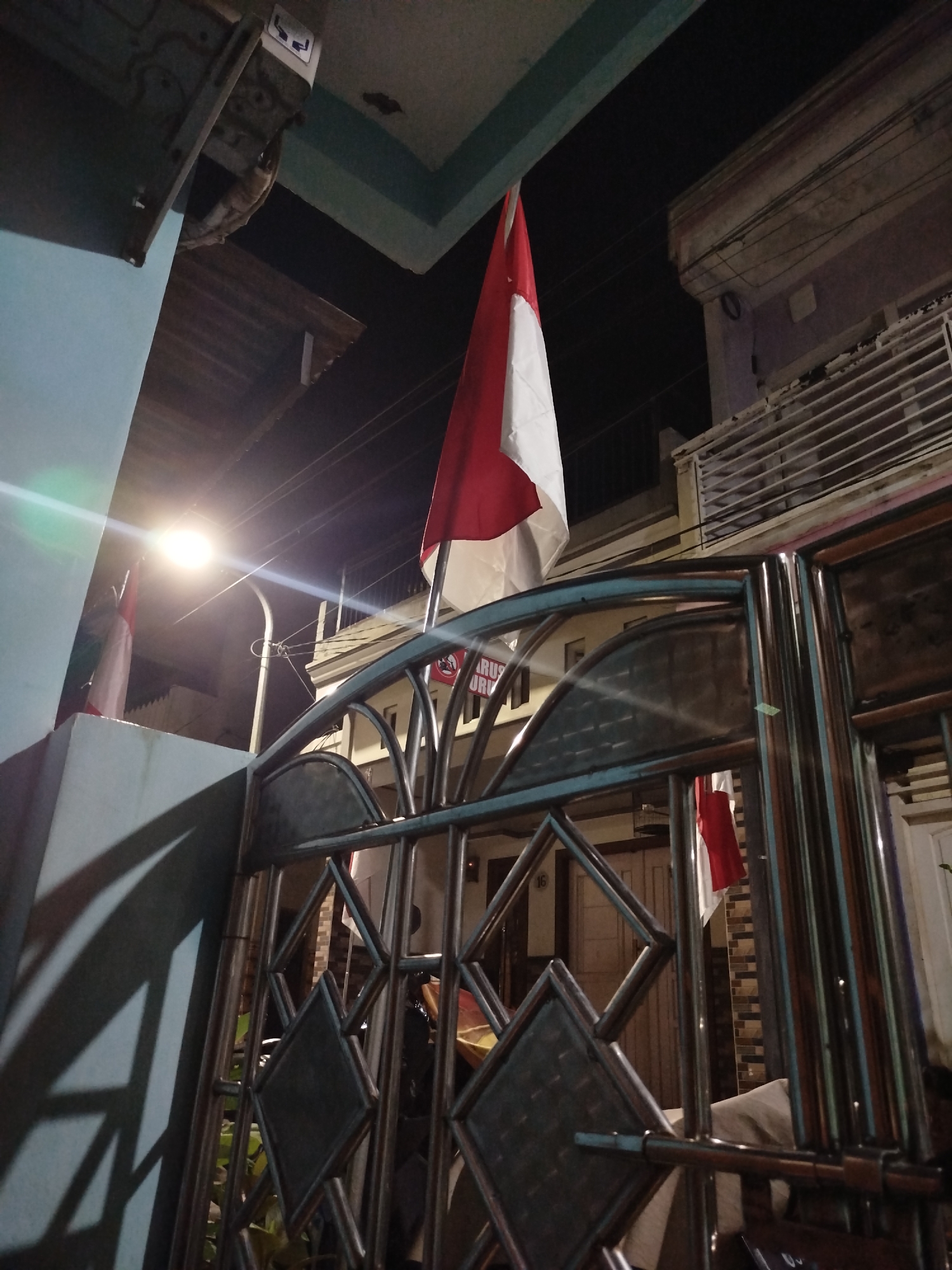 The image depicts a low-angle, nighttime view of a metallic gate in front of a building. A red and white flag, likely the Indonesian flag, is prominently displayed, hanging from a pole at the top left of the frame. A street light illuminates the scene, casting shadows on a light blue wall to the left of the gate. The gate itself is made of polished metal with diamond-shaped patterns. Behind the gate, glimpses of buildings with windows are visible, and another red and white flag can be seen further back. The overall environment suggests an urban setting, possibly residential, at night. The location is identified as Surabaya, Indonesia.