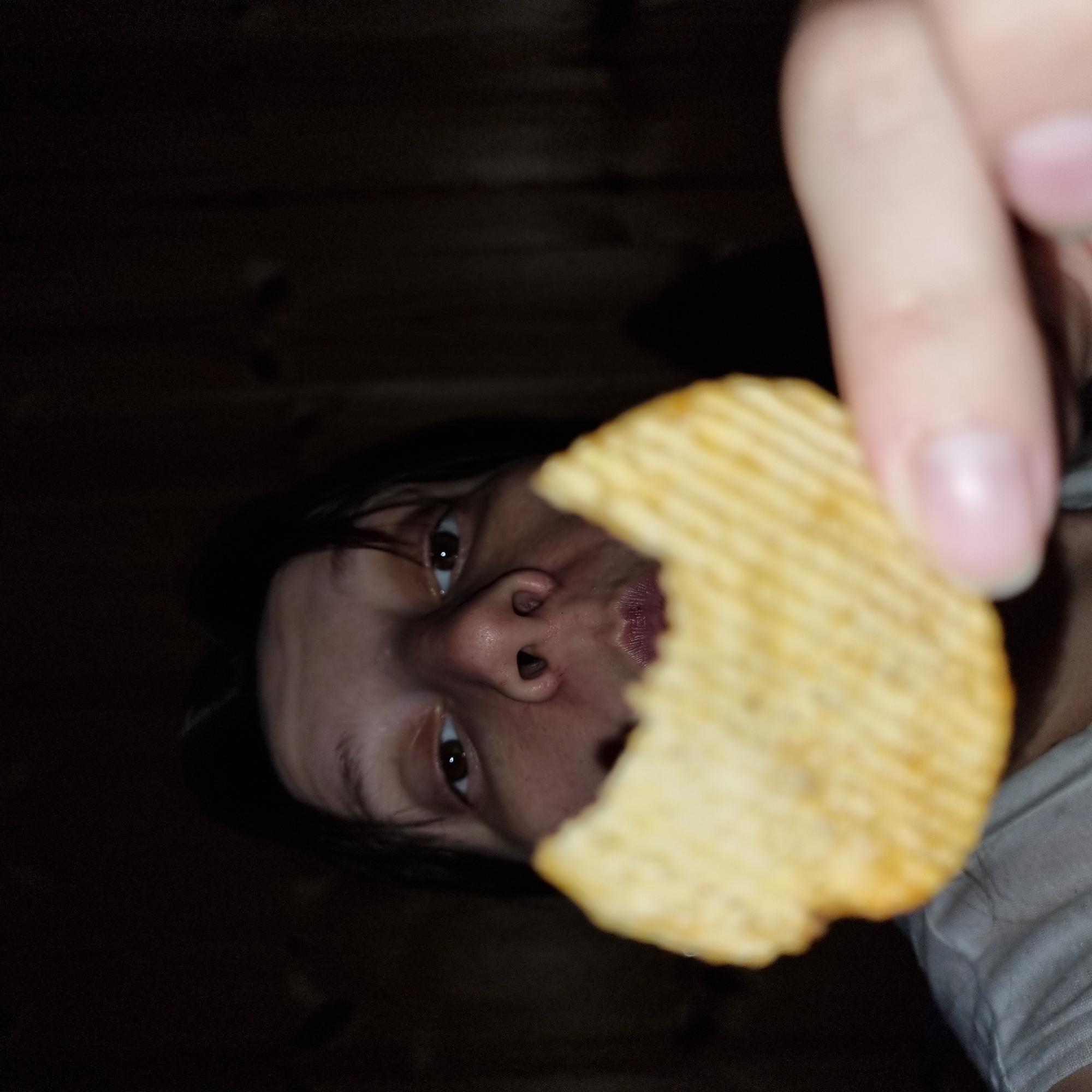 A close-up shot, taken from a low angle, shows a person holding a ridged potato chip close to their face. The person's face is partially obscured by the chip, with only their eyes, nose, and mouth visible. Their eyes are brown and looking directly at the camera. The person is wearing a grey t-shirt. The background is dark and out of focus, appearing to be wooden paneling, suggestive of an indoor setting. No specific details about the city of Dunnae-myeon, South Korea can be ascertained from the image.