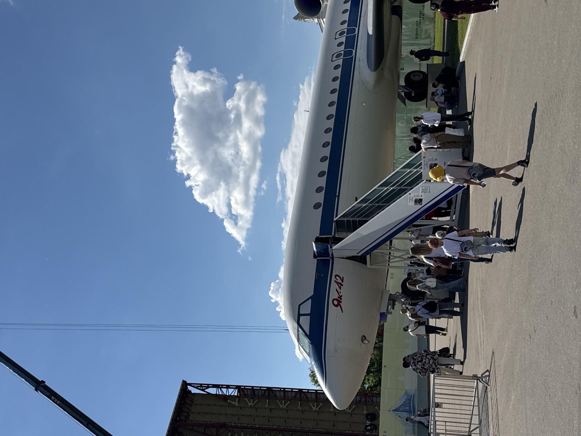 This vertically oriented image captures a bright, sunny outdoor scene featuring a large white and blue passenger aircraft, identified by the red Cyrillic lettering "Як-42" (Yak-42) on its nose. The aircraft, an Il-42, is a prominent static display, likely at an exhibition or museum in Khimki, Russia.
A metal boarding ramp extends from an open door on the aircraft's side, leading to a paved ground area. Numerous casually dressed individuals are gathered around the plane, some appearing to queue to enter or simply observing the impressive machinery. Their distinct, long shadows stretch across the asphalt, indicating bright daylight, likely late morning or early afternoon, under a clear blue sky dotted with scattered white cumulus clouds.
In the foreground, a person wearing a yellow hat and shorts walks away from the plane, their shadow sharply defined. To the left, parts of a large structure with scaffolding and a crane arm are visible against the sky, along with thin power lines. Behind the aircraft, a green sign with partially visible Cyrillic text, including phrases like "ВОДНОЕ ШОУ" (Water Show) and "30 МЕТРОВ" (30 meters), suggests other attractions or information nearby. A metal barrier or fence separates the crowd in some areas. The scene conveys a calm, engaging atmosphere, with people showing interest in the displayed aircraft.