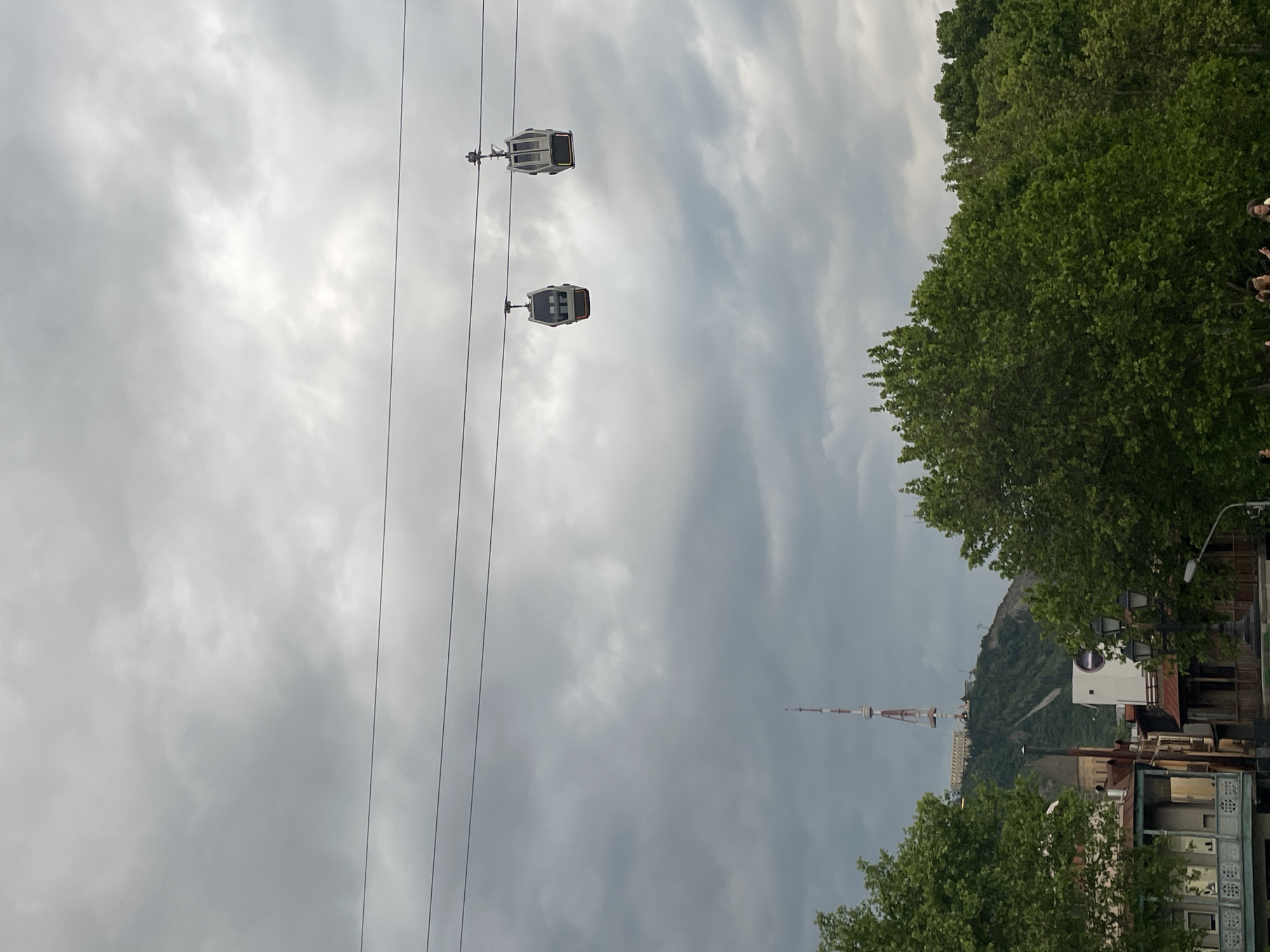 The image shows a cable car system in Tbilisi, Georgia, against a cloudy sky. Two gondola cars are suspended from cables. In the background, the Tbilisi TV Broadcasting Tower is visible on a hill, along with buildings and greenery. Trees line the right side and bottom of the image. The scene seems to be taken from a street-level perspective, looking upwards.
