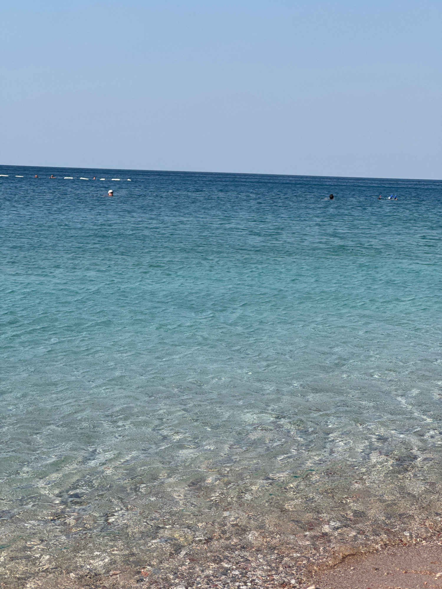 The image shows a clear, calm sea with a visible seabed in the foreground. The water transitions from a deep blue to a turquoise hue as it approaches the shore. Several people are visible in the water: one person wearing a white cap is swimming in the mid-ground, and two other individuals are further out, possibly snorkeling with blue fins visible. A line of white buoys stretches across the horizon. The sky is a pale, clear blue. The beach in the foreground is composed of small pebbles and sand.