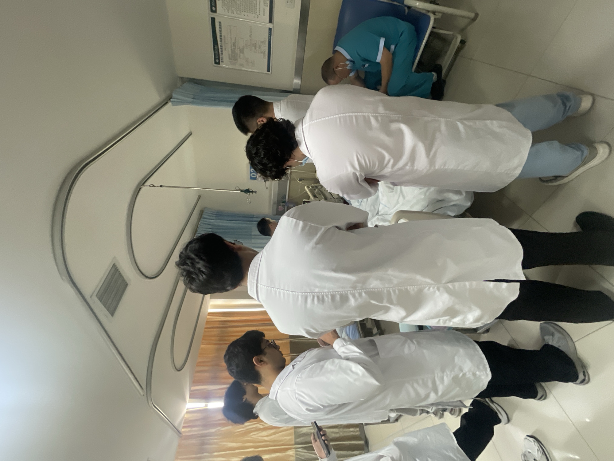 The image captures a scene inside what appears to be a hospital room, most likely in Tongliang District, China. Several people, mainly men wearing white lab coats, are gathered around a patient in a bed. One individual is wearing turquoise medical scrubs. Some are bent over, possibly examining or attending to the patient. The room features typical hospital furnishings, including a bed, medical equipment, and wall-mounted informational charts.