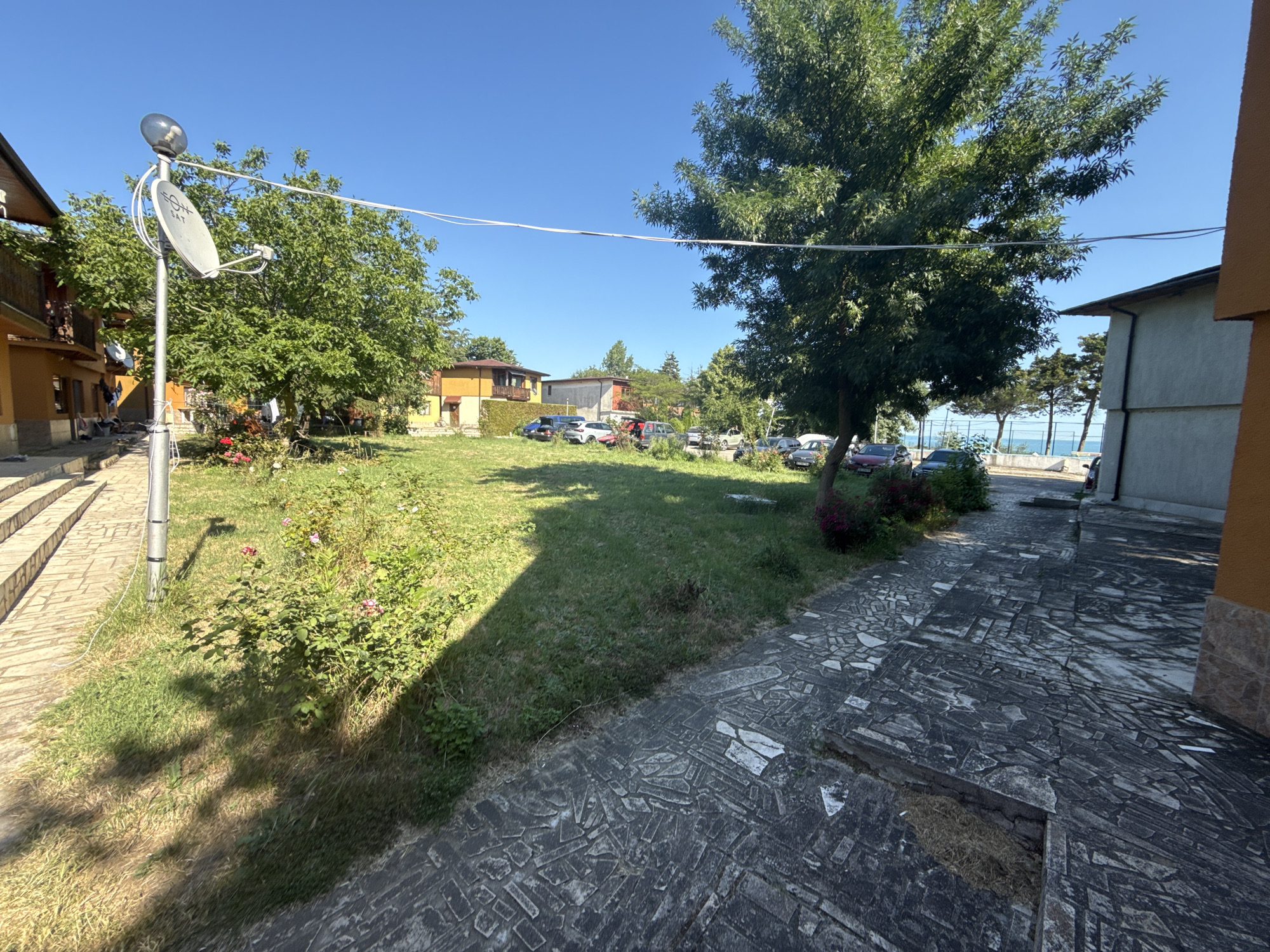 In Durankulak, Bulgaria, on a sunny day with a clear blue sky, a residential area is depicted. In the foreground, a stone-paved walkway leads to a grassy area with scattered bushes and flowers. A metal pole with a satellite dish and a light fixture stands to the left, partially casting a shadow on the grass. Behind the grass, several cars are parked in front of a row of houses and buildings. Some of the buildings have balconies and visible windows. Further in the background, trees line the horizon, with a glimpse of the sea and a distant fence suggesting proximity to the coast. The overall scene conveys a calm, everyday atmosphere in a coastal Bulgarian town.