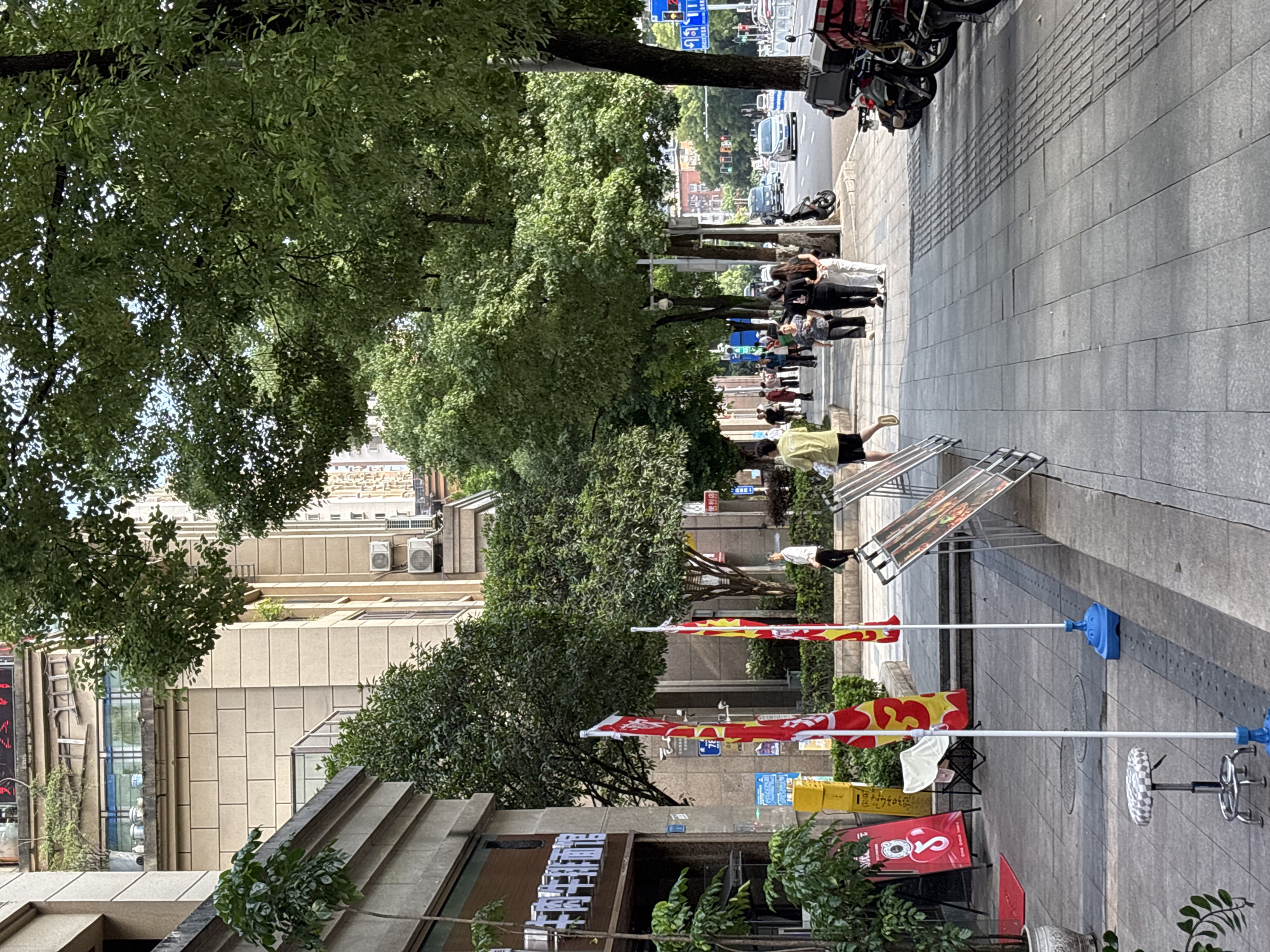 The image depicts a street scene in Yuzhong District, China. On the left side of the image, a building with beige tiled walls and several windows stands. In front of the building, there is a sidewalk with a few potted plants and red flags. Two people are visible walking on the sidewalk. Further down the street, several other people are walking, and some are seated on chairs. There are also several motorcycles parked along the side of the road. The street is lined with trees, and traffic can be seen in the distance, including cars and motorcycles. The overall scene suggests a commercial or urban environment with moderate pedestrian and vehicle activity.