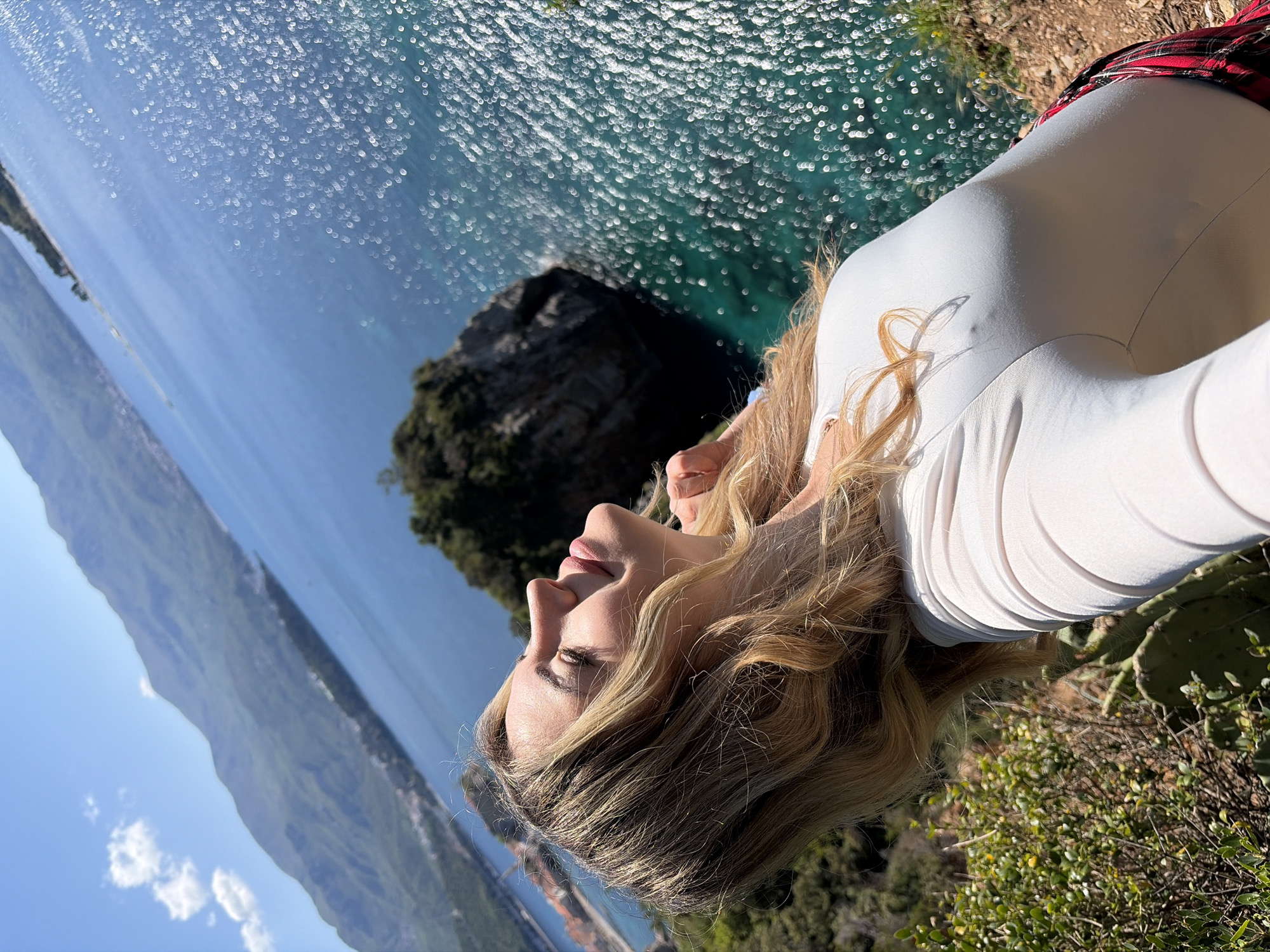 A woman poses in a scenic outdoor shot, seemingly at the edge of a cliff or high vantage point. The location is likely along the coast near Budva, Montenegro, as evidenced by the clear, turquoise water with light reflecting off the surface, a rocky island offshore, and a mountainous coastline in the distance. The woman is fair-skinned with blonde hair, wearing a white shirt and a red plaid skirt. She is standing among green vegetation, close to prickly pear cactus.