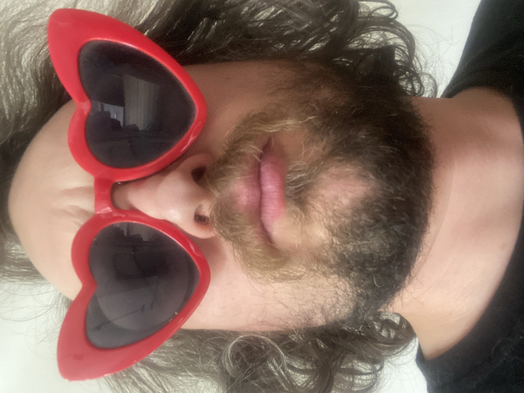 An individual with dark, curly hair and a beard is wearing red, heart-shaped sunglasses. The person's face is centered in the frame and appears to be angled upwards. The background is out of focus and appears to be a light-colored surface. The setting and location of the image in Rusanovo, Russia, are not discernible.