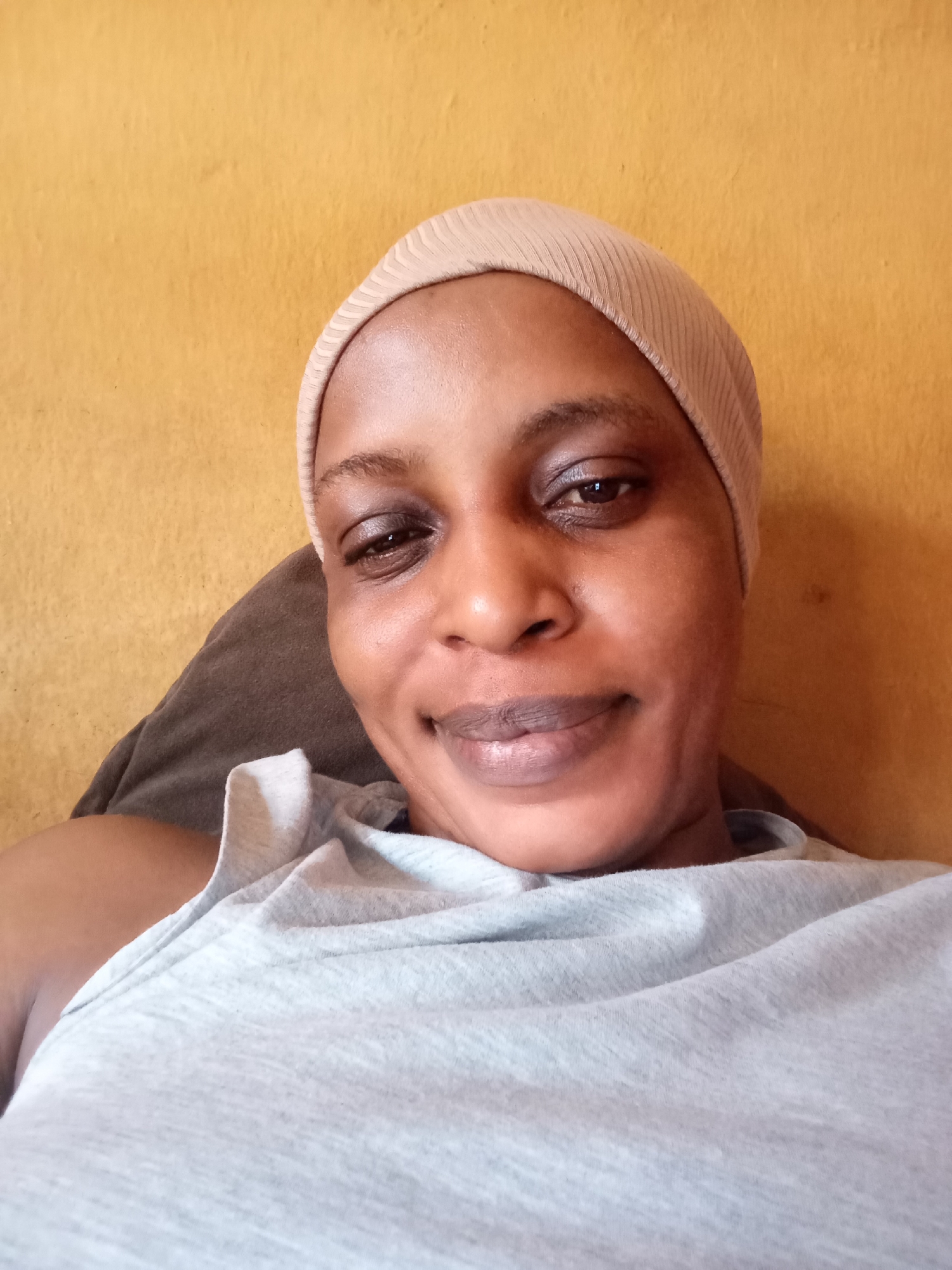 A medium shot captures a woman with dark skin, positioned against a mustard-yellow wall. She is wearing a beige knit cap and a light grey t-shirt. The woman is smiling slightly at the camera. A brown pillow supports her head. The location is in Narayi, Nigeria.