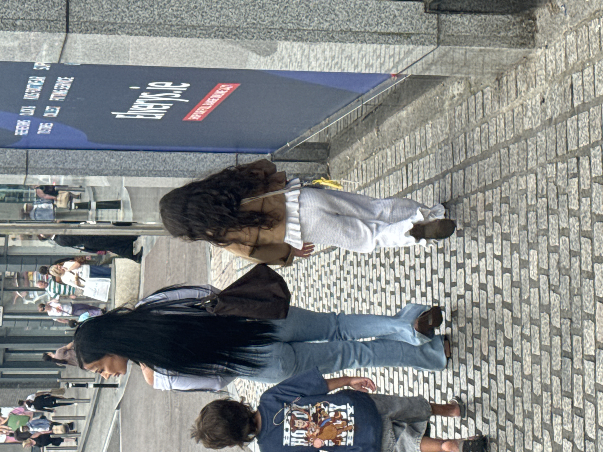 The image depicts a street scene in Waterford, Ireland, specifically the area near the Elvery's store on George's Street. A woman with long, dark hair walks away from the camera, wearing a white off-the-shoulder top and blue flared jeans. Beside her, a young girl with dark hair, also walking away, is dressed in a light brown jacket and white ruffled pants. To the left, a young boy in a blue t-shirt with a cartoon print walks ahead, wearing grey shorts. The group is walking on a cobblestone sidewalk, with a large blue Elvery's advertisement visible on the right side of the frame. The architecture suggests an urban environment, with buildings and a street in the background. The lighting indicates it is daytime, possibly overcast.